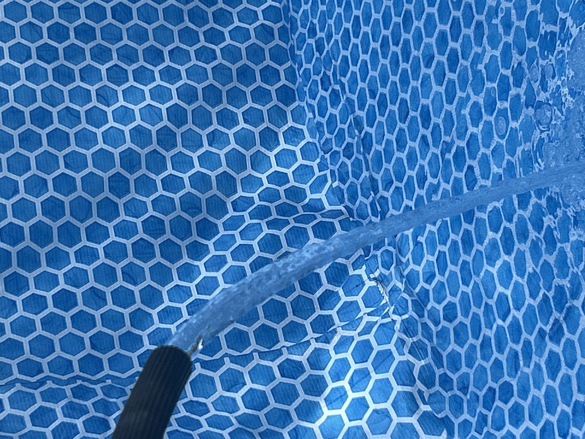 A close-up shot shows a stream of water being poured from a black hose onto a blue surface with a white hexagonal pattern. The water is a light blue and appears to be clear. The hexagonal pattern on the blue surface is slightly distorted in places due to the water. The scene is abstract, with the focus on the texture and movement of the water. The lighting is bright, suggesting it is daytime. The overall impression is one of a simple, everyday action captured in detail. There is no visible text in the image. The context of Prokhladny, Russia, does not provide specific visual cues within the image itself.