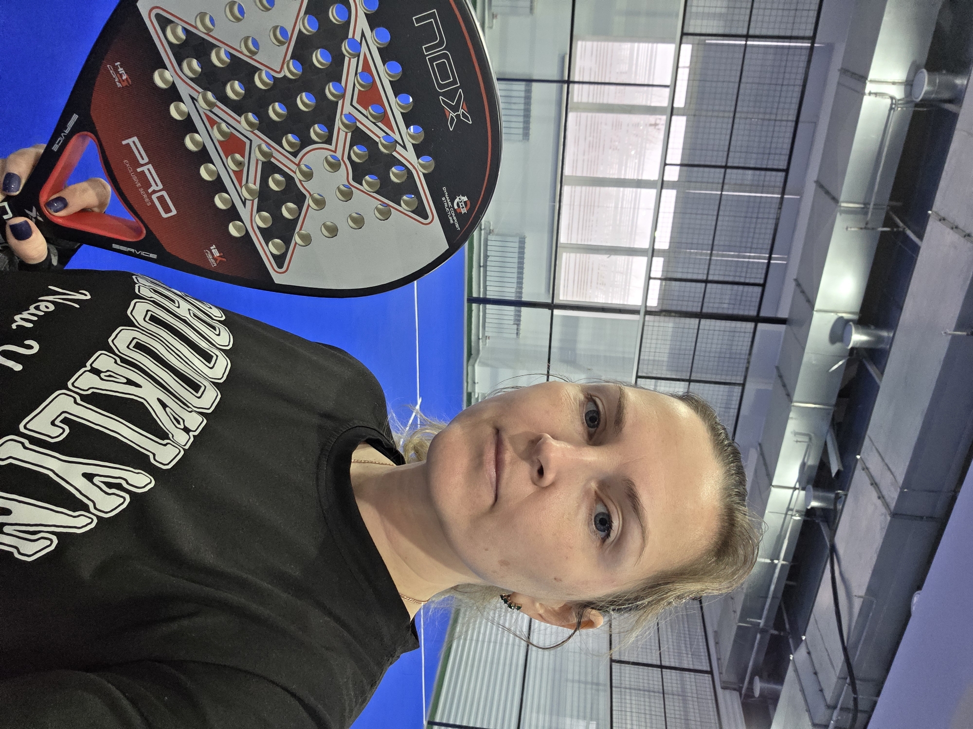 A woman takes a selfie on an indoor padel court in Chaikovsky, Russia. She has fair skin, light-colored hair, and blue eyes, looking directly at the camera with a neutral expression. She wears a black t-shirt with a white graphic. Her left hand, adorned with dark blue nail polish, holds a black, red, and white padel racket.
The padel racket is branded "NOX PRO EXCLUSIVE SERIES" and features text including "SERVICE," "12K CARBON," "HR3 CORE," and "DYNAMIC COMPOSIT STRUCTURE." The woman's t-shirt has the word "BOOKLY" in a prominent white font, with additional partially visible text suggesting "New York." The background shows the bright blue floor of the padel court. Behind her, large transparent or mesh panels indicate the court's boundaries and overhead structure, common in indoor padel facilities. Industrial-style ventilation ducts and lighting fixtures are visible on the ceiling, characteristic of a sports complex. The setting suggests she is either preparing for or has just finished playing padel.