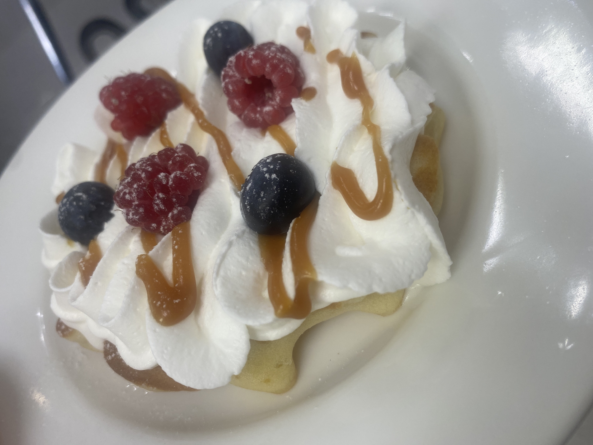 A close-up, overhead shot captures a single dessert item presented on a white, circular plate. The dessert consists of a light-colored, pancake-like base generously topped with swirls of white whipped cream. Drizzled over the whipped cream is a caramel-colored sauce, creating an abstract pattern. Scattered across the dessert are fresh berries, including bright red raspberries and dark blue blueberries. A light dusting of powdered sugar is visible on the whipped cream and the berries. The background is out of focus, but appears to be a clean, possibly commercial kitchen environment with faint hints of stainless steel equipment. The image is lit from above, highlighting the textures and colors of the dessert. There are no people or animals present in the image. The dessert is the sole focus. There is no information in the image that suggests a specific location in Kolomna, Russia.