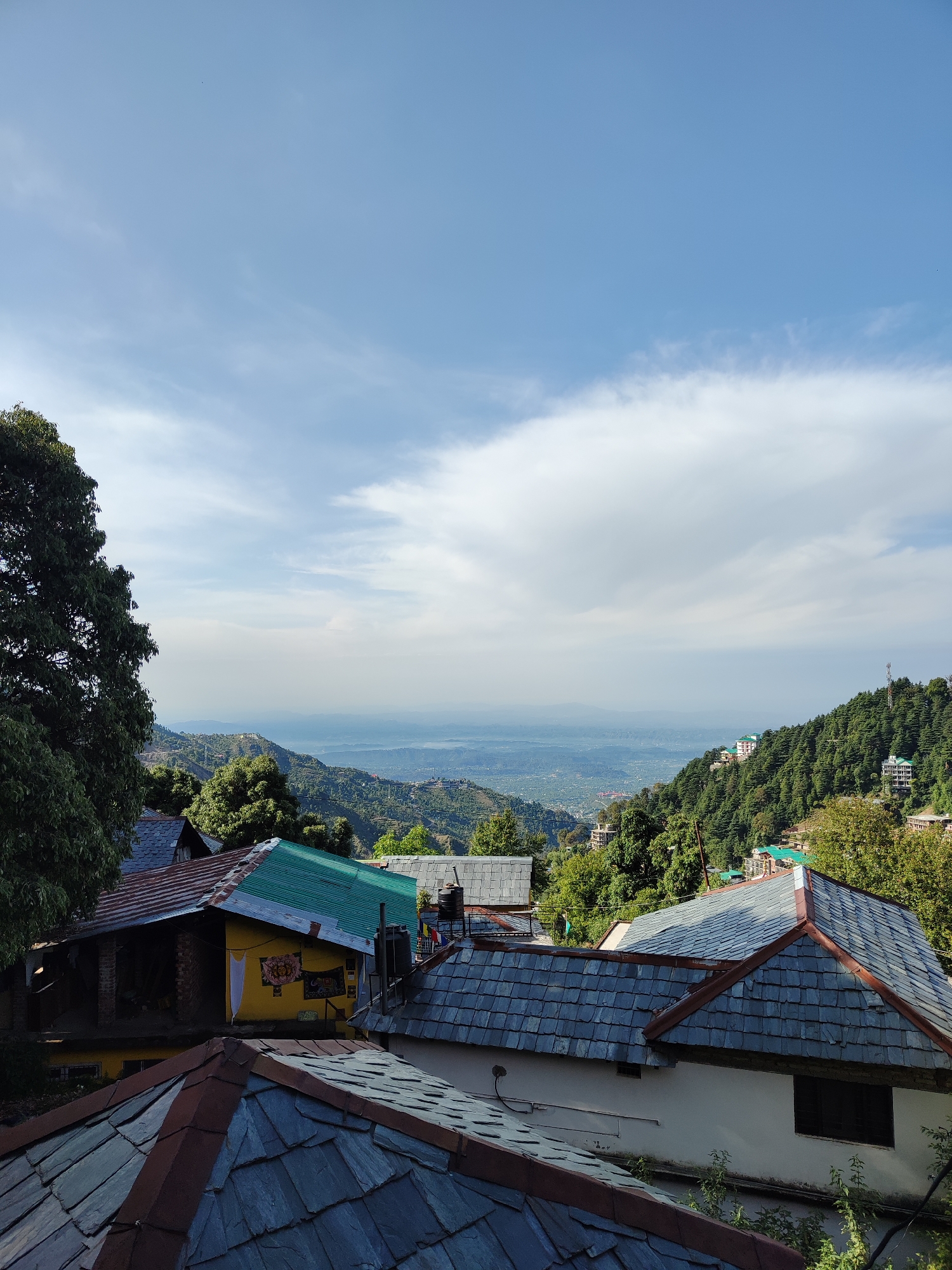 The image shows a daytime view of a mountainous landscape dotted with houses, likely taken from an elevated position in the city of Haini, India. The foreground features rooftops constructed from slate tiles, varying in color from gray to brown. Behind these structures, a yellow house is visible with what appear to be tapestries or wall hangings. Lush green trees are scattered throughout the scene, covering the surrounding hillsides. In the distance, there is a hazy valley, with additional hills and mountains beyond. The sky is predominantly blue with patches of white clouds. The overall lighting is bright and natural, indicating a sunny day.