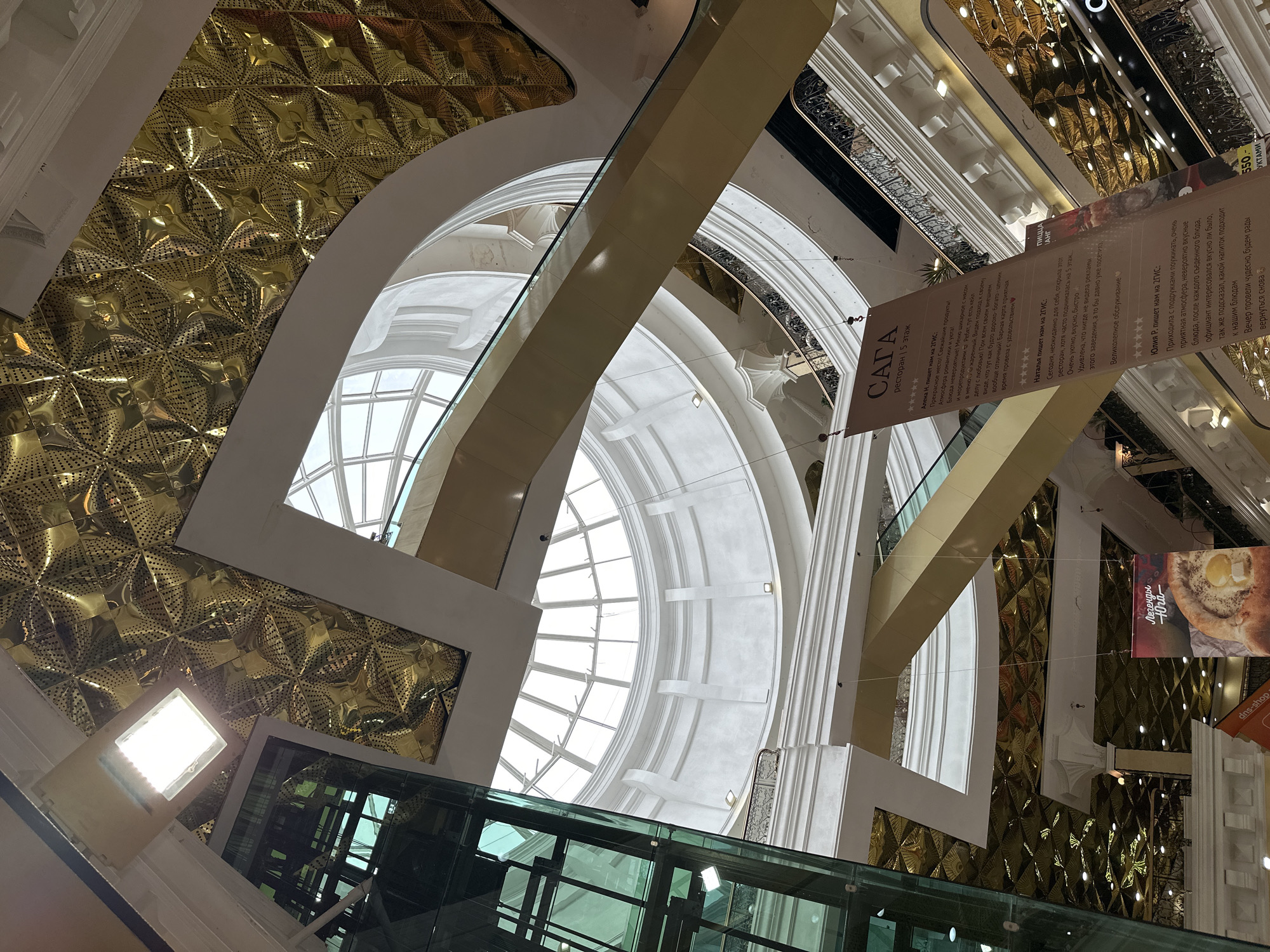 The image captures an upward perspective within the opulent, multi-story interior of a commercial complex, likely a shopping mall, located in Yekaterinburg, Russia. The scene is dominated by grand, classical-inspired architecture, featuring vast arched windows that allow abundant natural light to illuminate the space, indicating daytime. The design incorporates extensive golden, textured panels with a distinct star-like or floral pattern, juxtaposed with smooth white and beige curvilinear structural elements and arches.
Connecting the various levels, a long escalator with a glass balustrade ascends diagonally, and a glass-enclosed elevator shaft is visible at the bottom left. Ornate railings adorn the balconies of the upper floors, contributing to the luxurious aesthetic. Artificial light fixtures are also present, complementing the daylight.
Several vertical banners, all in Russian, are suspended within the space, advertising various establishments. A prominent banner promotes "САГА ресторан 5 этаж" (SAGA restaurant 5th floor) and showcases glowing customer reviews praising the restaurant's "Прекрасное место! Свежайшие продукты! Атмосфера романтики и уюта! Блюда невероятные!" (Wonderful place! Freshest products! Atmosphere of romance and comfort! Incredible dishes!). Other visible text includes "ПИЦЦА АНГ" (PIZZA ANG), "Легенды Юга" (Legends of the South) next to an image of khachapuri, and a partial "dns-shop" sign for an electronics retailer. The image focuses on the architectural grandeur and commercial offerings, with no people explicitly visible.