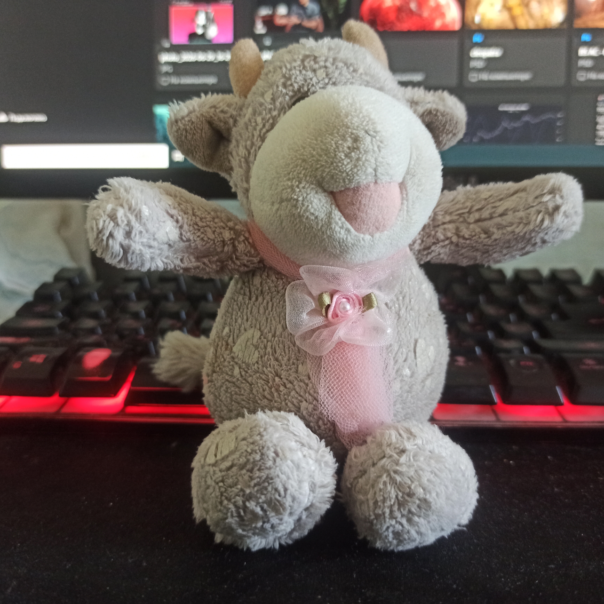A stuffed toy cow sits on a black mousepad in front of a computer keyboard with red backlighting and a computer monitor displaying various content. The cow is light gray with a white face and pink accents. It wears a pink scarf with a small pink bow adorned with a pearl. The cow's arms are outstretched. The scene is indoor and appears to be a home or office environment. There is no information available that would allow for the identification of its location in Nizhny Novgorod, Russia.