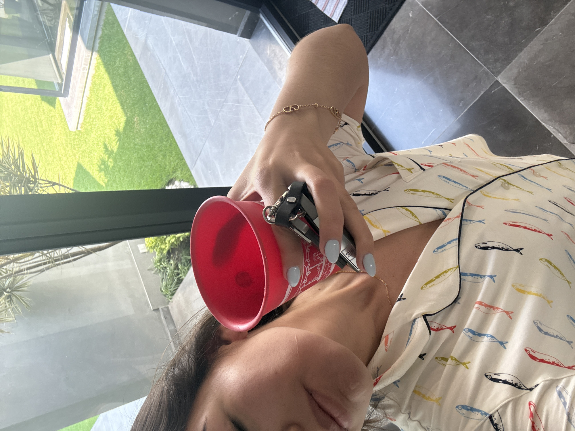 The image captures a person, likely a woman, from an overhead, diagonal perspective, holding a red cup and a set of keys. She wears a white pajama-style shirt patterned with colorful fish in red, blue, yellow, and black, accented with dark piping. Her left hand, adorned with light blue nail polish, holds the red cup and a black key fob. A gold chain bracelet with a double 'V' or 'VV' logo is visible on her wrist, and she wears a delicate gold necklace.
The setting appears to be indoors, near a large glass window or door that looks out onto a sunny outdoor area. The outdoor view features a bright green lawn with shadows, indicating bright daylight, possibly morning or afternoon. Adjacent to the lawn is a light-colored paved or tiled area and some green foliage. The indoor floor consists of large grey square tiles. The scene suggests a relaxed moment at home, potentially in San Bernardino Tlaxcalancingo, Mexico. No discernible text is visible in the image.