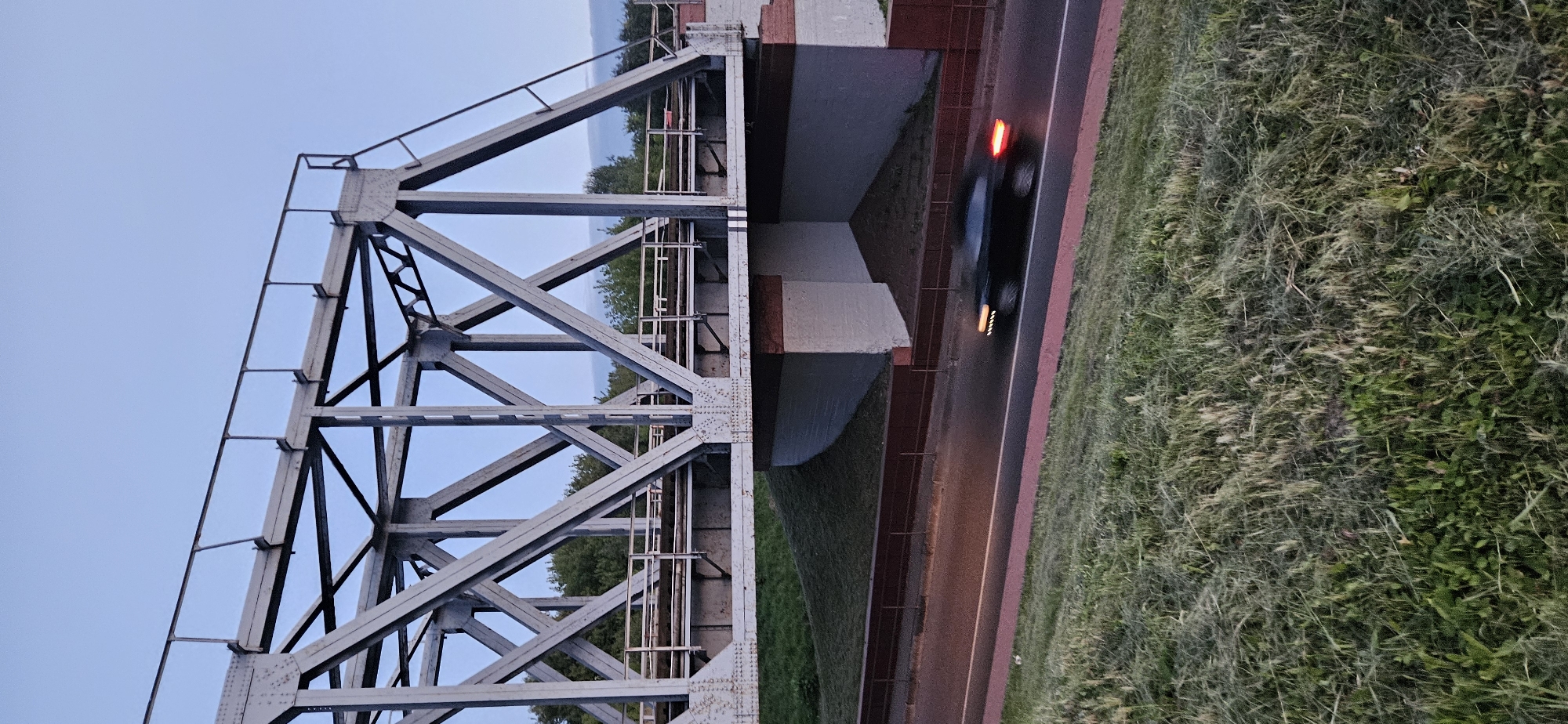This image shows a metal truss bridge over a road. The bridge has a complex structure of beams and supports, painted a light gray. There are train tracks on top of the bridge. Below the bridge, a black car is driving on the road, captured with motion blur, indicating it is moving. The car's headlights and taillights are visible. The road has a red line marking on its side. The scene is surrounded by green grass on a sloping embankment. Trees are visible in the background, behind the bridge. The sky is a pale blue, suggesting it is either early morning or late evening. There is no visible indication of the location being Vitsebsk, Belarus in the image itself.