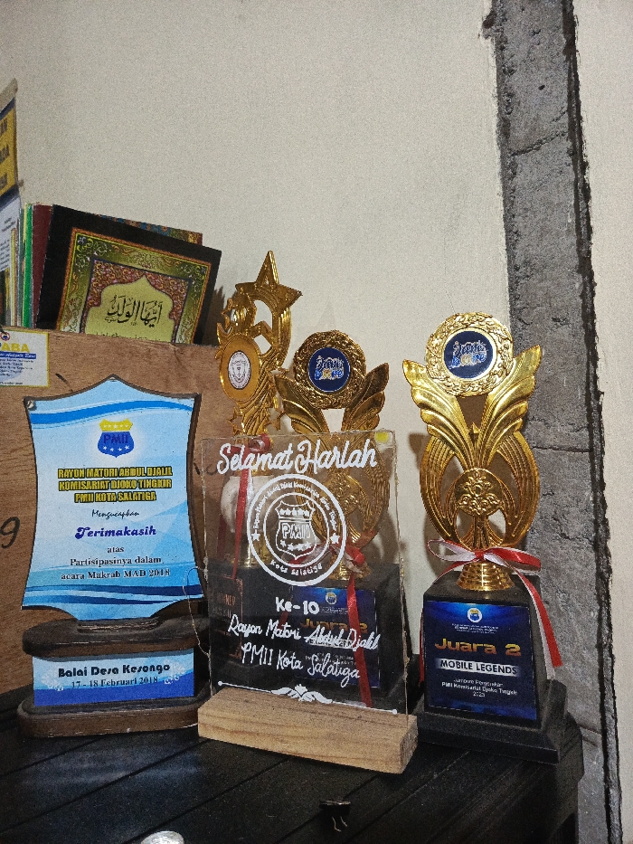 A collection of awards, plaques, and books is displayed on a dark wooden surface against a light-colored wall, with a visible concrete seam or pillar on the right. The setting is likely indoors, in Tuntang, Indonesia.
On the left, several books are stacked, one prominently showing Arabic text that reads "أيها الولد" (Ayyuhal Walad). Below this, a blue and white shield-shaped plaque expresses thanks from "RAYON MATORI ABDUL DJALIL KOMISARIAT DIOKO TINGKOR PMII KOTA SALATIGA" for participation in "acara Makrab MAD 2018," held at "Balai Desa Kesongo" (Kesongo Village Hall) on "17 - 18 Februari 2018."
In the center, a clear acrylic plaque with a wooden base features the text "Selamat Harlah" (Happy Anniversary/Birthday), celebrating the "Ke-10 Rayon Matori Abdul Djalil PMII Kota Salatiga" (10th Anniversary of the Rayon Matori Abdul Djalil branch of the Indonesian Islamic Student Movement in Salatiga City). Behind it is a golden trophy with a star design.
To the right, two more golden trophies are visible. One has a distinctive leaf-like design and a blue circular emblem. The furthest right trophy, decorated with a red and white ribbon, signifies "Juara 2 MOBILE LEGENDS" (2nd Place in Mobile Legends), awarded to "PMII Komisariat Djoko Tingkir." The display highlights achievements in organizational events and competitive gaming.