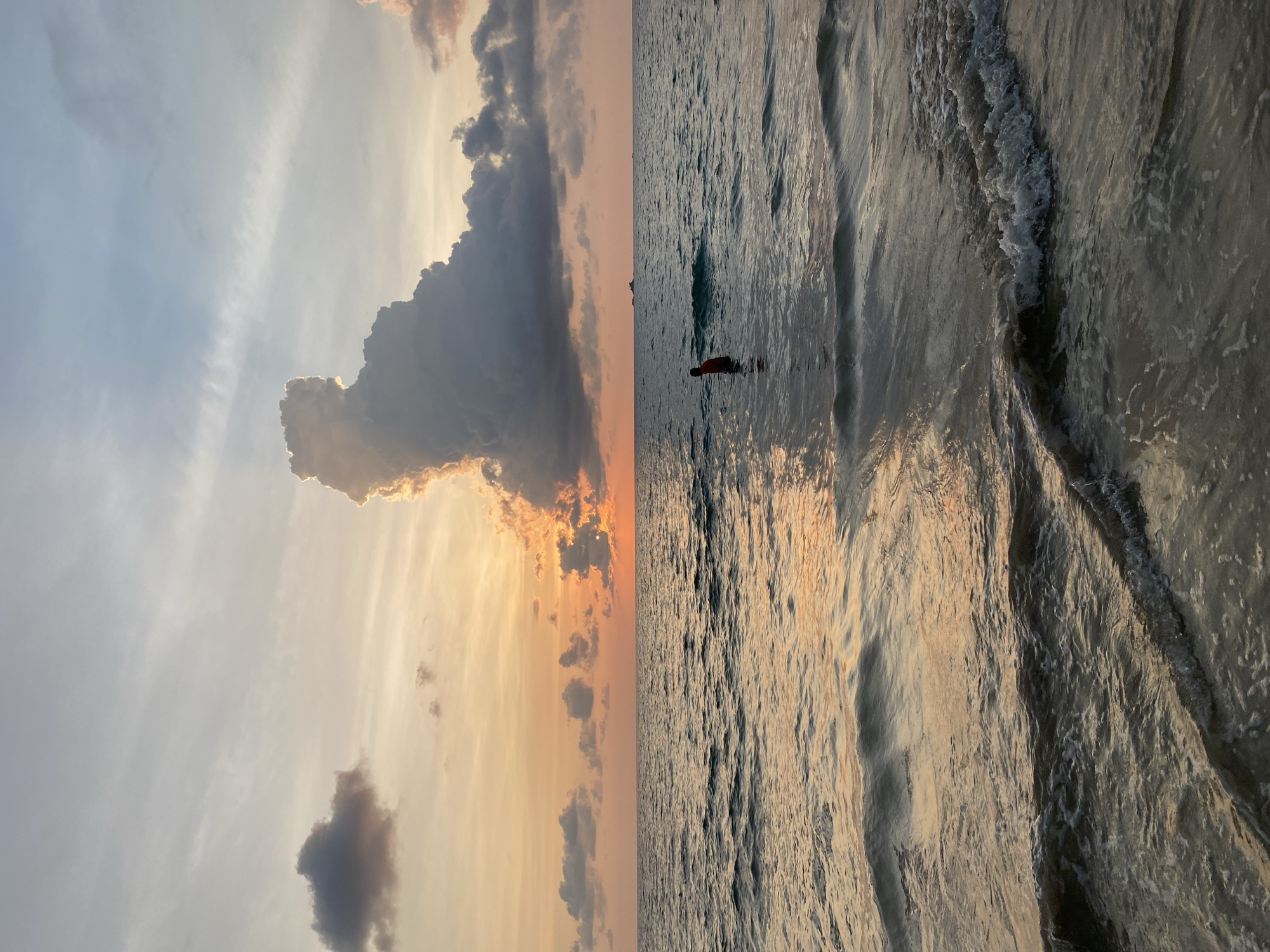 The image depicts a tranquil coastal scene, likely a beach or ocean, at either sunrise or sunset in Choeng Thale, Thailand. The horizon line, when viewed correctly, separates the expansive sky from the reflective water.
The upper portion of the image is dominated by a dramatic sky. Large, dark, and voluminous clouds are prominent, especially on the right side, where they are brilliantly backlit by the sun's golden and orange glow. This warm light also spreads across lighter, wispy clouds and parts of the sky, creating a vibrant palette of yellows, oranges, and soft greys. Towards the left, the sky transitions to a lighter blue-grey with more diffused cloud cover.
The lower half of the image showcases the surface of the water, which appears calm with gentle waves and ripples. The water beautifully reflects the warm, golden light from the sky, creating shimmering highlights across its texture. In the mid-distance, a lone figure can be seen standing or wading in the shallow water. The person, appearing small against the vastness, is dressed in what looks like a reddish-orange top, adding a small point of interest to the expansive scene.
The overall atmosphere is serene and contemplative, highlighting the natural beauty of the coastal environment during a picturesque time of day. No visible text is present in the media.