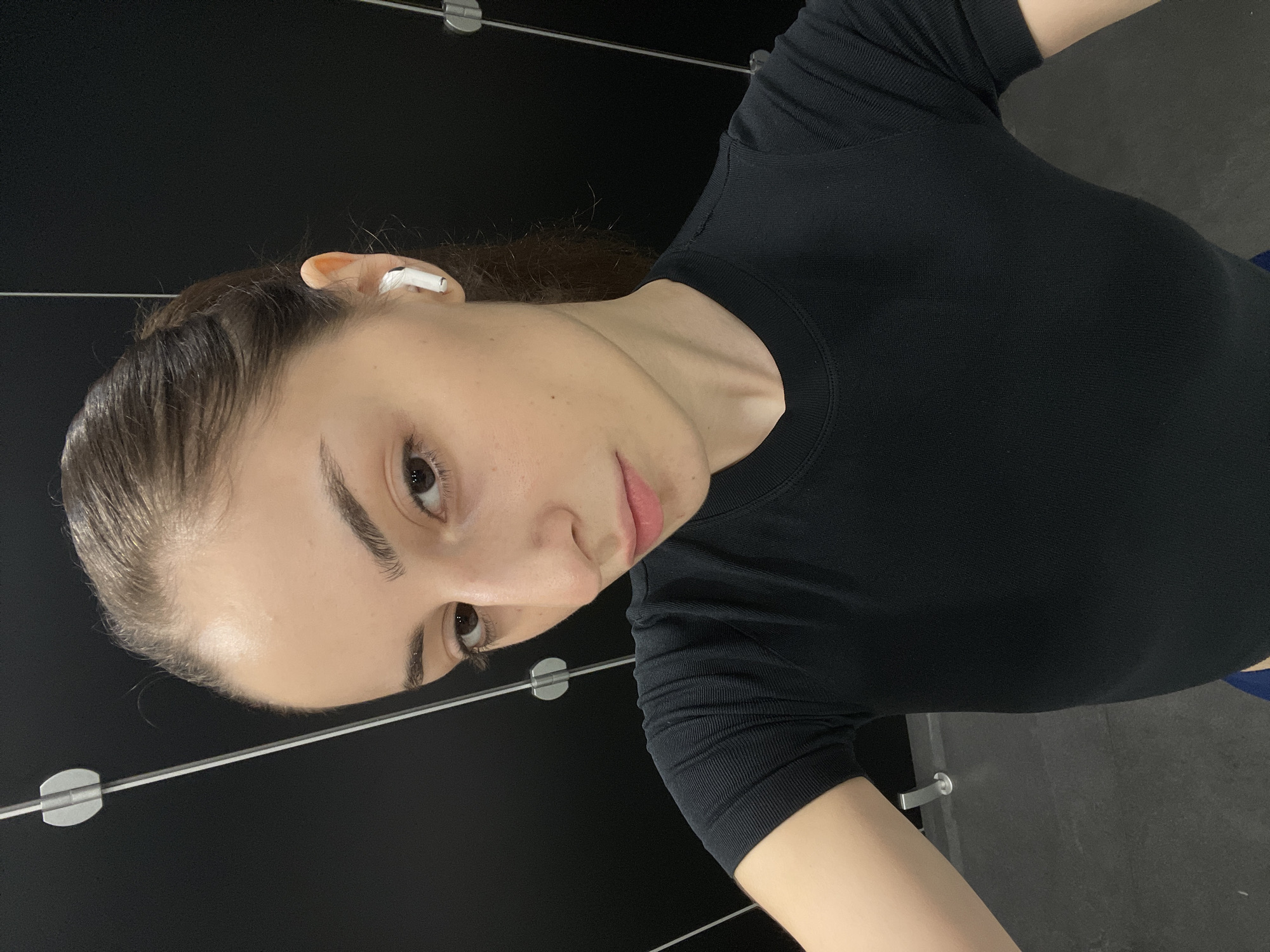 A young woman with brown hair pulled back in a ponytail is taking a selfie in what appears to be a locker room. She is wearing a black short-sleeved shirt and has white earbuds in her ears. The background is a dark wall with vertical metal dividers. The flooring is dark grey. There are no visible indicators of her location in Moscow, Russia.