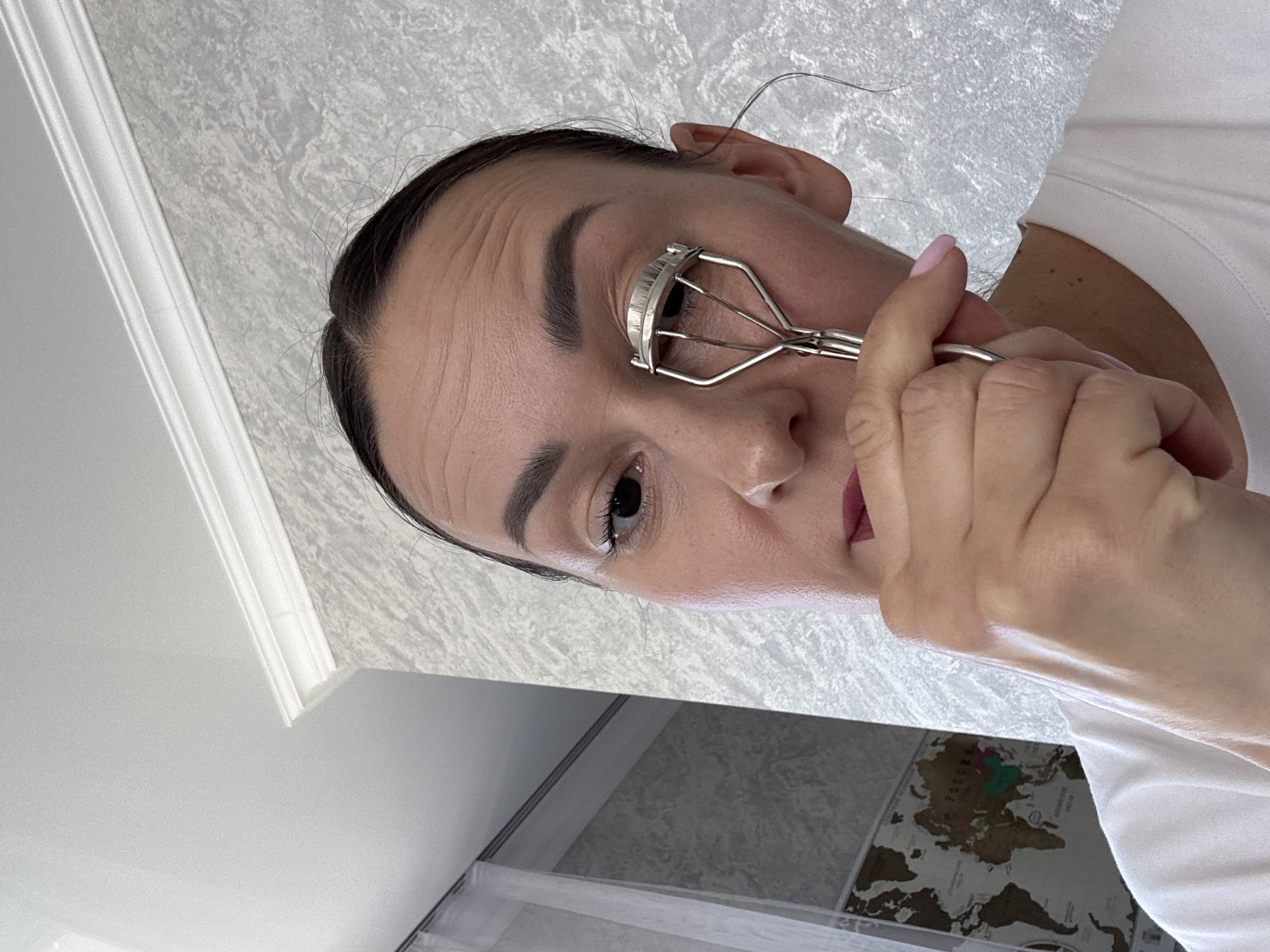 The image, taken in Sochi, Russia, captures a close-up, angled view of a woman applying makeup. She is seen from the chest up, with her dark hair pulled back, revealing her forehead which shows some horizontal lines. Her left eye is in focus, where a silver metal eyelash curler is positioned, indicating she is in the process of curling her lashes. Her left hand, with light pink painted nails, holds the curler. She appears to be wearing subtle eye makeup and possibly lipstick. She is dressed in a plain white t-shirt. The background features a light-colored, textured wall or ceiling with white architectural molding on the upper left. In the lower right corner, a framed scratch-off world map is visible, showing various countries, some of which appear to be colored in. The lighting suggests it is daytime. The woman's expression is neutral and focused on her task. No discernible text is clearly legible on the world map.