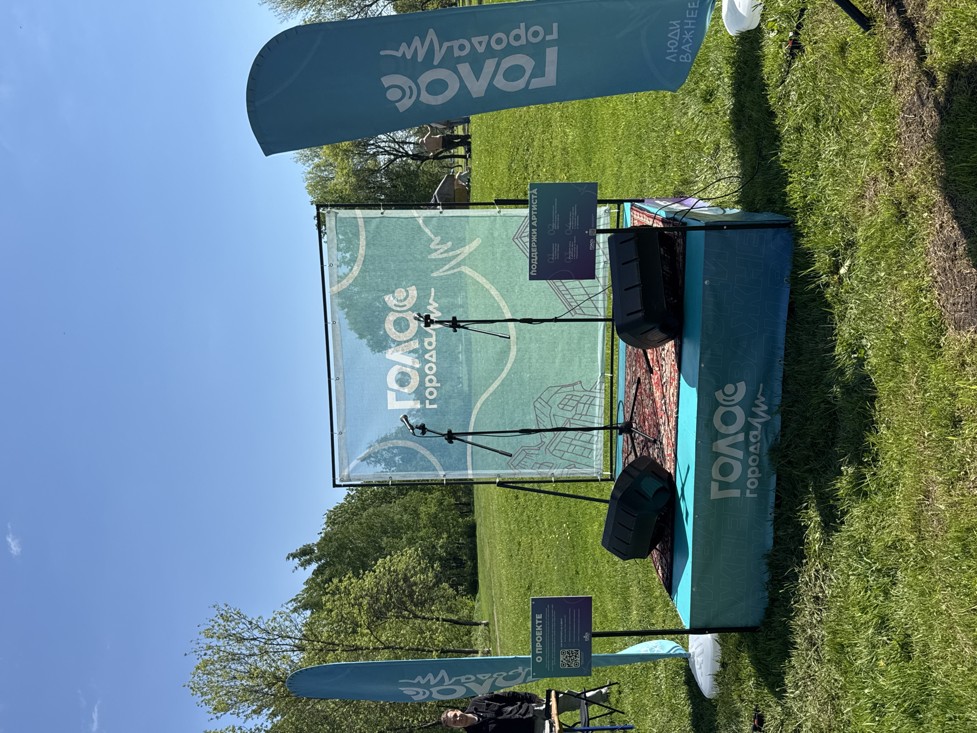 Here is a description of the image:
The image shows an outdoor event or setup, likely a small stage for performances. The backdrop features the word "ГОЛОС" (Voice) with what appears to be an outline of a city. Two microphones stand on the stage. Speakers are placed on the sides of the stage. Banners are positioned on either side, also with "ГОЛОС" written on them, and some text on the banners including the phrase "люди важнее". A sign saying "О ПРОЕКТЕ" is also visible, along with a sign that says "ПОДДЕРЖИ АРТИСТА". The environment appears to be a grassy area, possibly a park, with trees in the background under a clear blue sky. A man is visible sitting in a chair in front of the stage. The location appears to be in Unknown, Unknown.