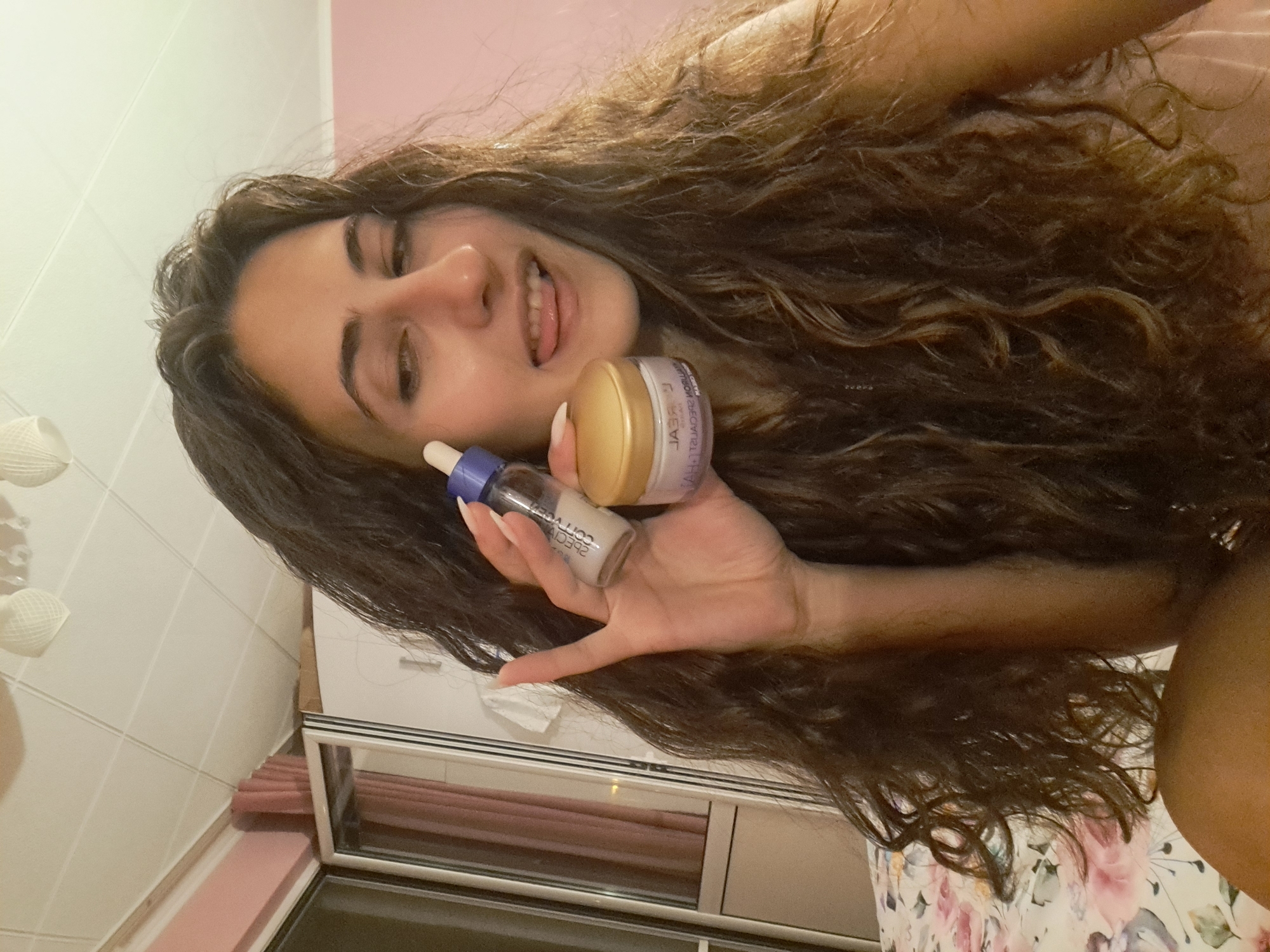A young woman with long, dark curly hair is shown indoors, holding two skincare products. She is looking at the camera with her tongue sticking out playfully. The room appears to be a bedroom, with a bed in the foreground covered with a floral patterned duvet. In the background, there is a closet with mirrored doors and a ceiling with white light fixtures. The wall behind the woman is pink. The scene suggests a casual and relaxed setting, possibly in a home. The image does not provide any specific geographical markers for the city of Negovan, Bulgaria, beyond the general indoor setting.
