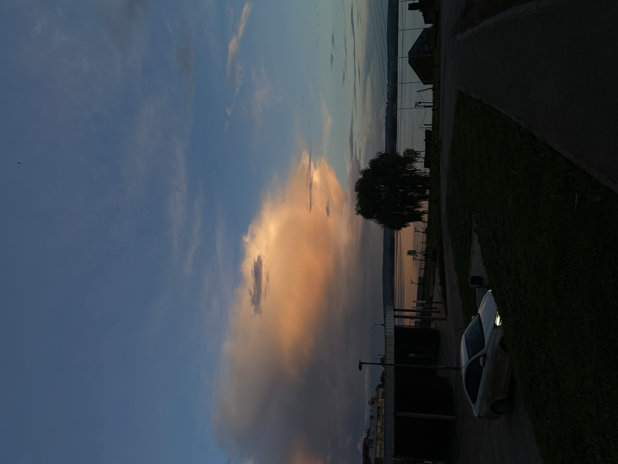 This image captures a tranquil twilight scene over a large body of water, likely the Votkinsk Reservoir in Votkinsk, Russia. The sky dominates the upper portion, showcasing a dramatic contrast between the deep blue color on the left and a prominent cloud formation on the right, vividly illuminated by the warm, soft hues of orange and pink, suggesting either sunrise or sunset. Wispy clouds are scattered across the blue expanse.
The large body of water occupies the mid-ground, reflecting the subdued colors of the sky, appearing dark blue-grey with subtle hints of the warm cloud colors. On the immediate shoreline, a dark, dense deciduous tree stands silhouetted against the water and sky. A paved path or road runs parallel to the water's edge, bordered by dark green grass. A white car is partially visible on this paved area, with its headlights or parking lights illuminated, reinforcing the dim light conditions. In the distance, across the water, a faint, dark outline of a distant shore or hills can be discerned. Along the near shore, various dark structures, possibly small buildings or elements of an embankment, are scattered. A yellow triangular road sign, likely indicating a pedestrian crossing, is partially visible near the water's edge in one of the close-up views, though no specific text is legible. A small dark speck, possibly a bird, is visible against the blue sky in the upper left. The overall scene is peaceful and devoid of human activity, capturing the quiet beauty of dusk or dawn.