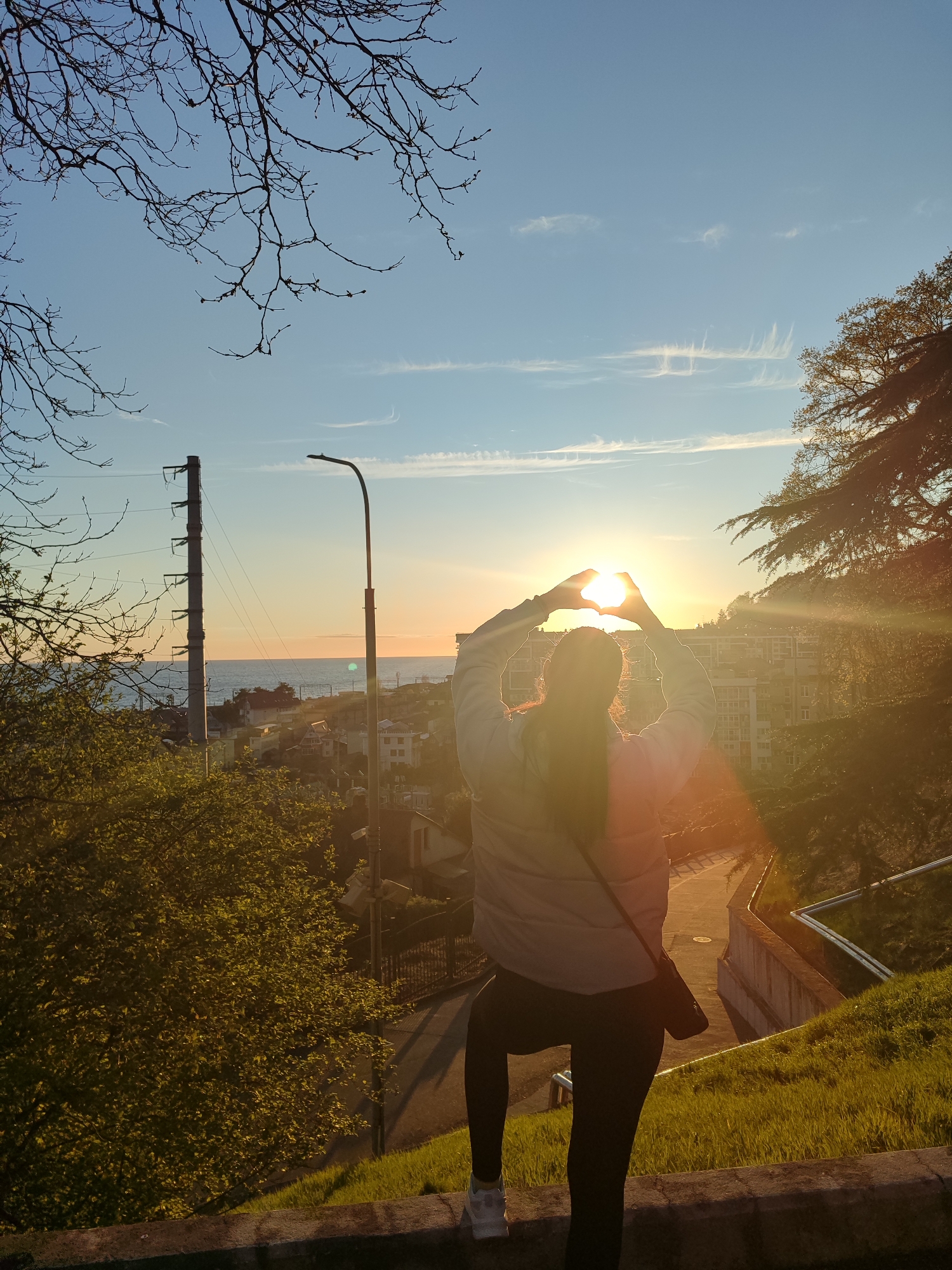 A woman stands facing away from the camera, with her arms raised to form a heart shape around the setting sun, in the city of Unknown, Unknown. She is wearing a light-colored jacket, dark leggings, and white sneakers, with a dark purse slung across her body. The sun is positioned centrally in the frame, casting a golden glow across the scene. To the left, there is a tall electrical tower, sparse tree branches, and green foliage. A street lamp stands between the tower and the woman. Buildings are visible in the background, to the right, along with a large tree casting long shadows. The scene takes place on a grassy hillside with a concrete ledge. A pathway leads down the hill to the right. The sky above is a gradient of blue and orange hues.