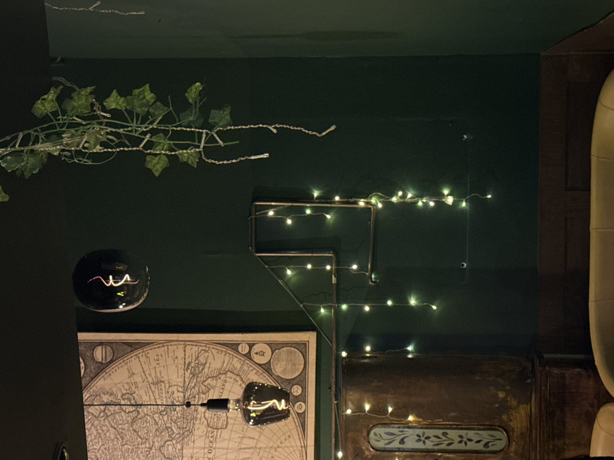 An interior scene in the city of Unknown, Unknown, presents a dimly lit space characterized by dark green walls. A large, framed vintage-style world map is mounted on the left wall. Below the map, a decorative Edison-style filament bulb in a dark, teardrop-shaped glass shade hangs from the ceiling. To the right of the map, a dark, spherical object containing an illuminated, twisted filament is affixed to the green wall. Below this, a structure of copper-toned pipes forms an angular shape on the wall, draped with multiple strands of warm white LED string lights that extend vertically downwards. In the upper right, artificial green ivy vines are interwoven with additional string lights, descending from the ceiling. The lower left reveals a section of a dark wooden cabinet or panel featuring a light-colored, stylized floral inlay. At the bottom edge, portions of two light-colored, upholstered seating units are visible. The overall illumination is low and warm, creating a subdued atmosphere. No individuals are present.