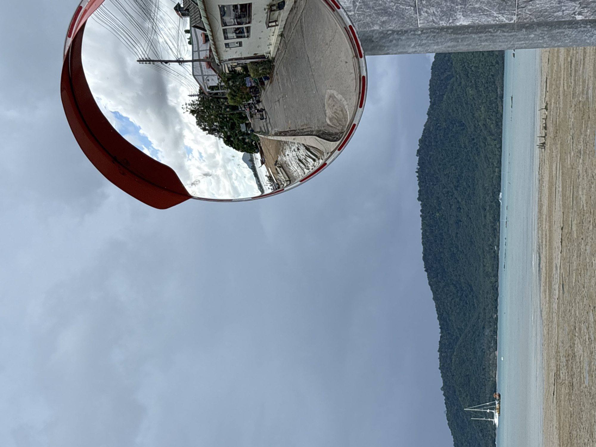 In Rawai, Thailand, a convex traffic mirror reflects a coastal road lined with buildings and lush trees. The reflection shows a paved road with a slight incline, a small motor scooter, and several parked motorcycles. A white building with windows and a bench is visible on the right side of the reflected road. In the distance, a sailboat with white sails is moored on a calm, turquoise sea. The sea is bordered by a sandy beach and a densely forested hill that occupies the background. The sky is overcast with gray clouds, creating a diffused light. The mirror itself is framed in red and white, mounted on a metal pole.