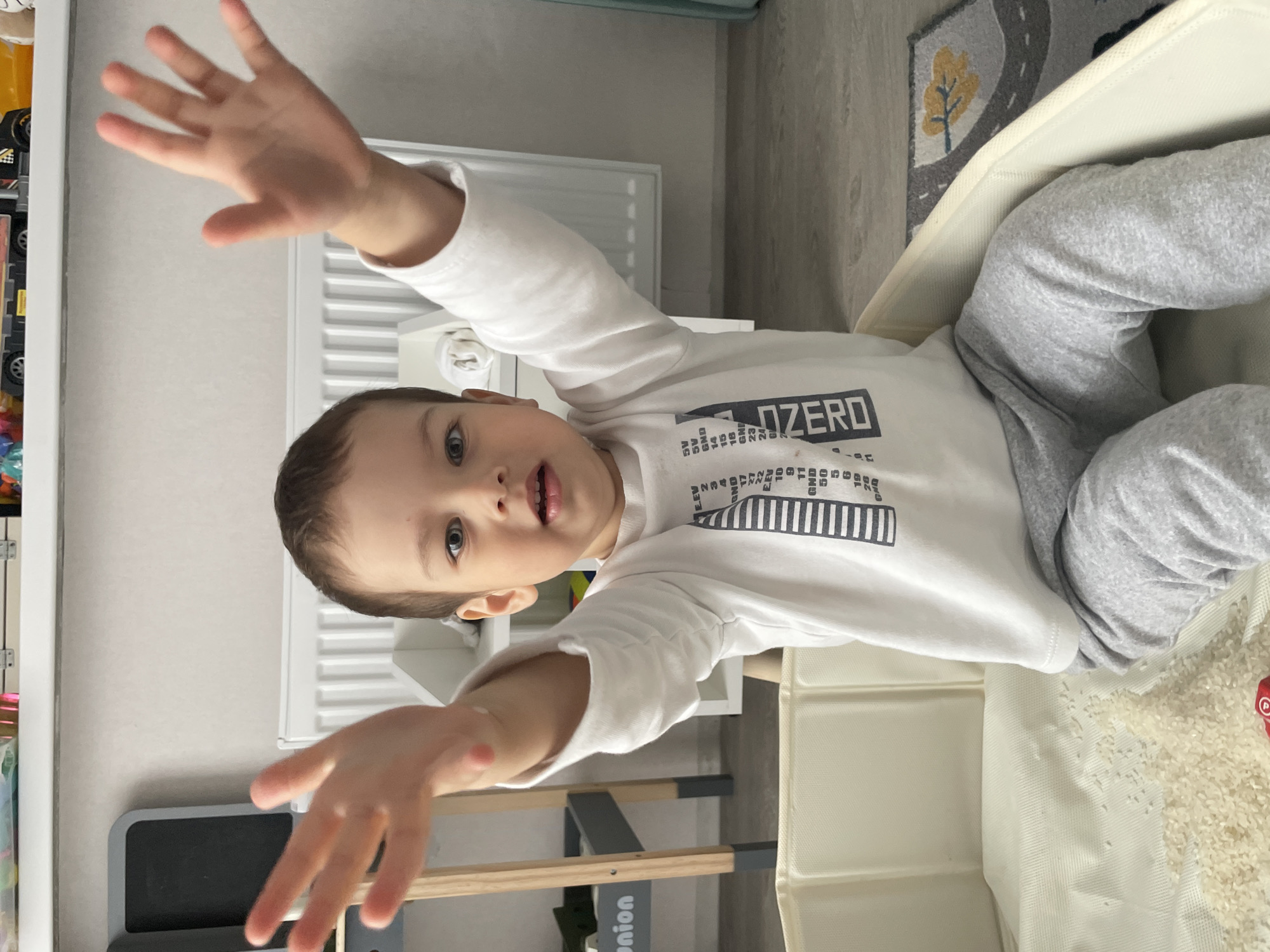 A young child is positioned in the center of the frame, partially inside a white, rectangular container filled with rice. The child is wearing a long-sleeved white shirt with a graphic print that includes the word "ZERO" and gray sweatpants. Their arms are extended towards the camera, with open hands.
Behind the child, there is a radiator against a wall, and to the left, a shelf with toys. A gray and white child's rug with a road pattern is visible at the top right of the image.