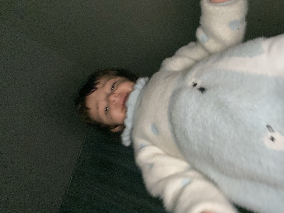 A baby in a light blue and white, fluffy, footed pajama lies on a dark surface. The pajama has a dotted pattern. The baby faces the viewer, eyes open. The baby is positioned at an angle, head toward the upper left and feet toward the lower right corner of the frame. The lighting is dim.