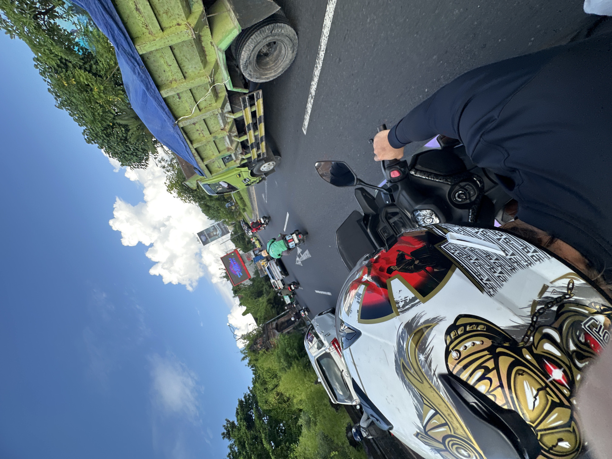 The image depicts a person riding a motorcycle on a road in Kuta, Indonesia. The view is from the rider's perspective, showing a helmet with an elaborate design predominantly in white, gold, black and red. The rider is wearing a long-sleeved dark top and holding the handlebars. Traffic is visible ahead, including a green dump truck to the left and other motorcycles and cars. A large billboard is situated on the left side of the road. The sky above is blue with scattered white clouds. Lush green trees and vegetation line the roadside.
