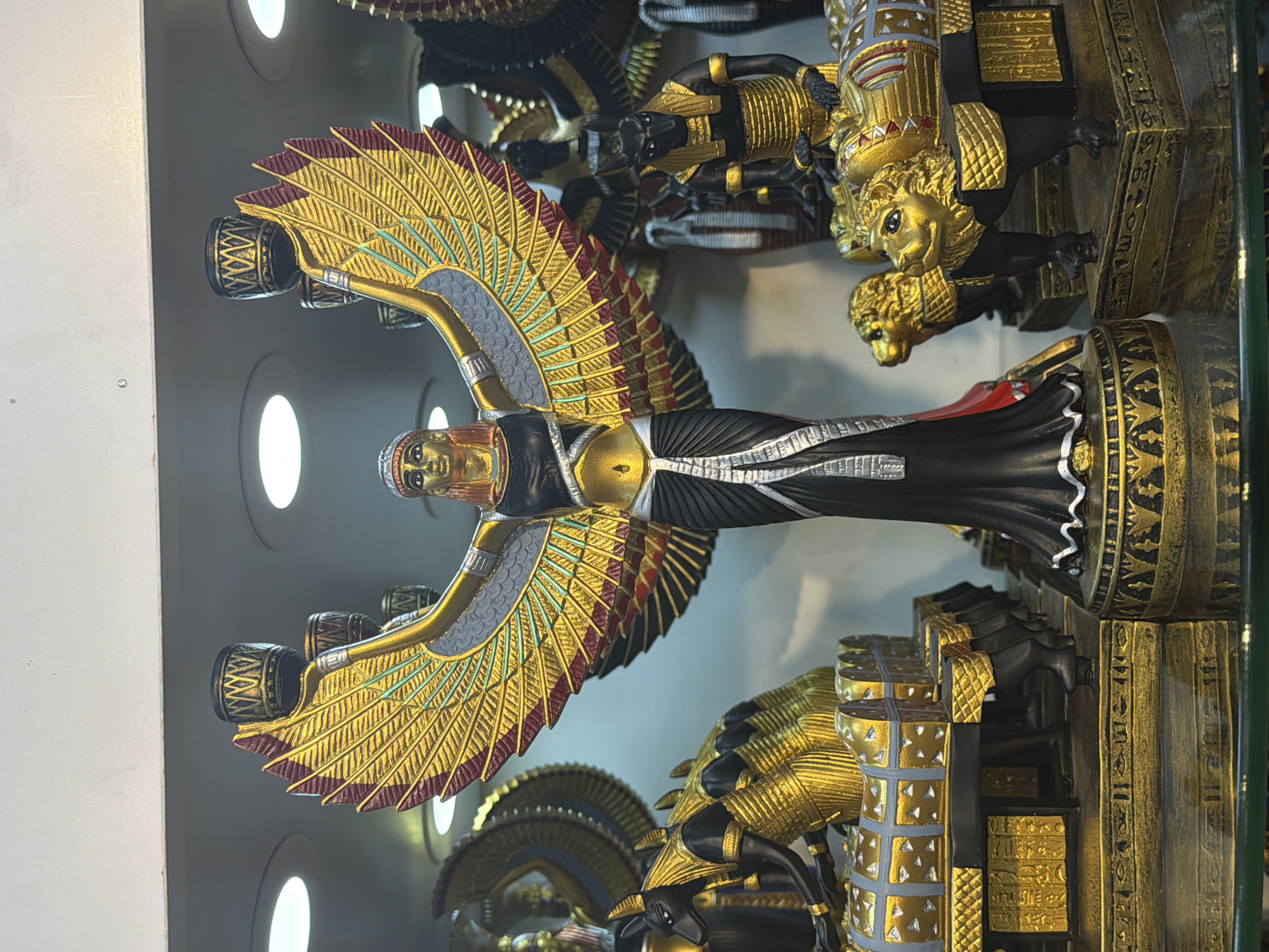 The image displays a collection of Egyptian-themed decorative figurines, likely for sale in a shop. The central figure is a winged goddess, possibly Isis or Nephthys, depicted with large, golden wings outstretched, holding two cylindrical objects at the top. She wears a black dress with a white sash and a golden headdress. To the left and right of the central figure are other figurines. On the left, there are several Anubis figures, characterized by their jackal heads and golden armor, seated behind elaborate, golden sarcophagi or chests adorned with hieroglyphs and decorative patterns. On the right, partially visible, is a golden lion-like creature with a regal mane, also part of a decorative structure. The figurines are placed on shelves with a reflective surface at the bottom, suggesting a display case in a retail environment. The background is out of focus, showing more similar decorative items and lighting fixtures. The overall impression is a display of souvenirs or artistic representations of ancient Egyptian mythology. The location is identified as Sharm El Sheikh, Egypt, implying a tourist-oriented retail setting.