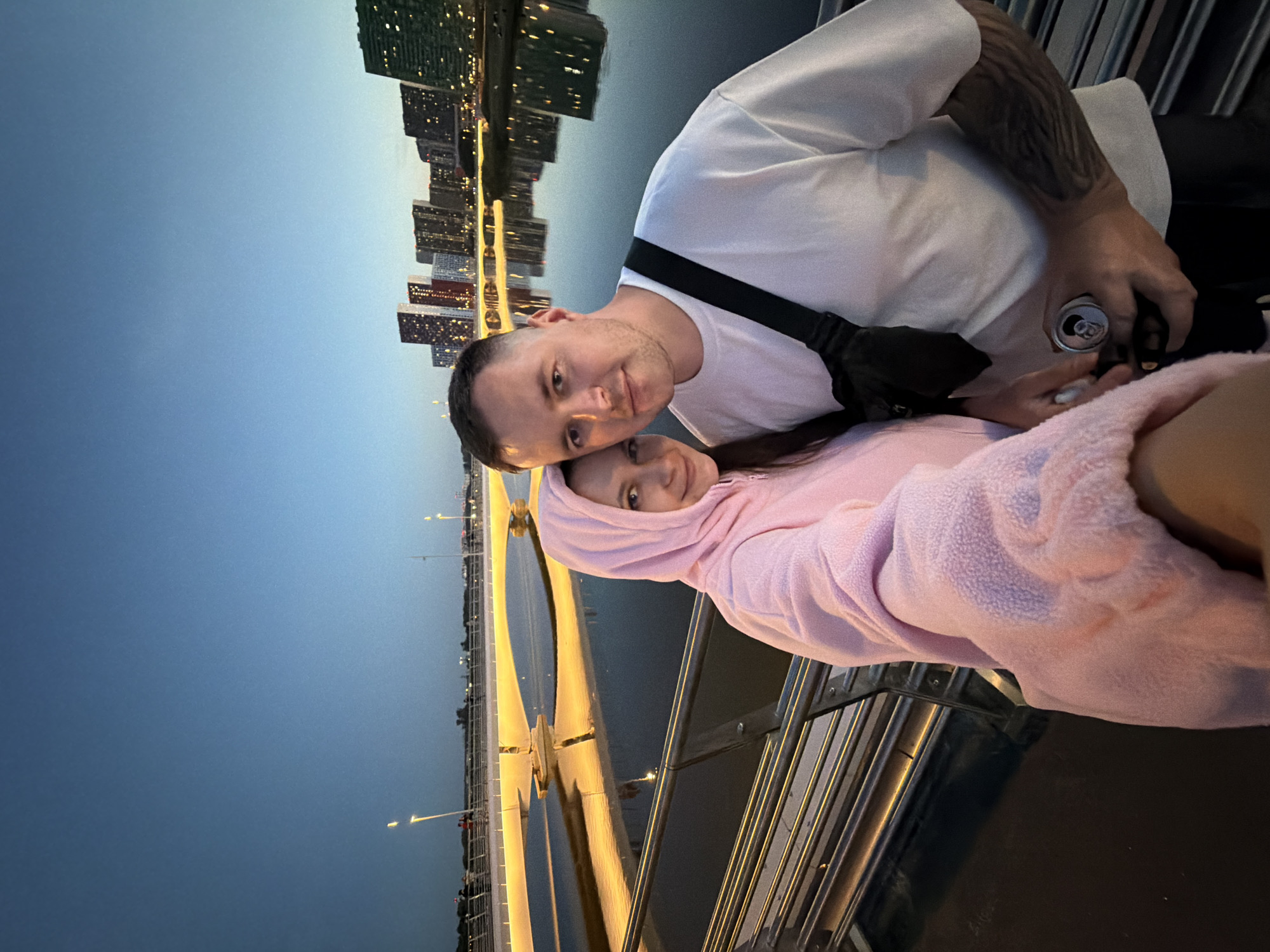 The image captures a selfie of a man and a woman on a modern pedestrian bridge at twilight in Moscow, Russia. In the foreground, a multi-bar metal railing runs horizontally. The woman, partially visible on the left, wears a pink hooded fleece jacket and is holding the camera. To her right, a man with short dark hair, a white t-shirt, and a black cross-body bag looks directly at the camera with a neutral expression. He has a dark tattoo on his left forearm and holds an open silver beverage can in his right hand.
The background features a calm body of water (likely the Moskva River) reflecting the golden-yellow lights of the contemporary bridge structure. Further in the distance, across the water, a cluster of modern, high-rise buildings, characteristic of the Moscow International Business Center (Moscow City), stands against a deep blue evening sky. Some windows in the skyscrapers are illuminated. Other blurred figures are visible on the bridge in the middle distance. The scene is illuminated by the artificial lights of the bridge and distant buildings under a clear, darkening sky.