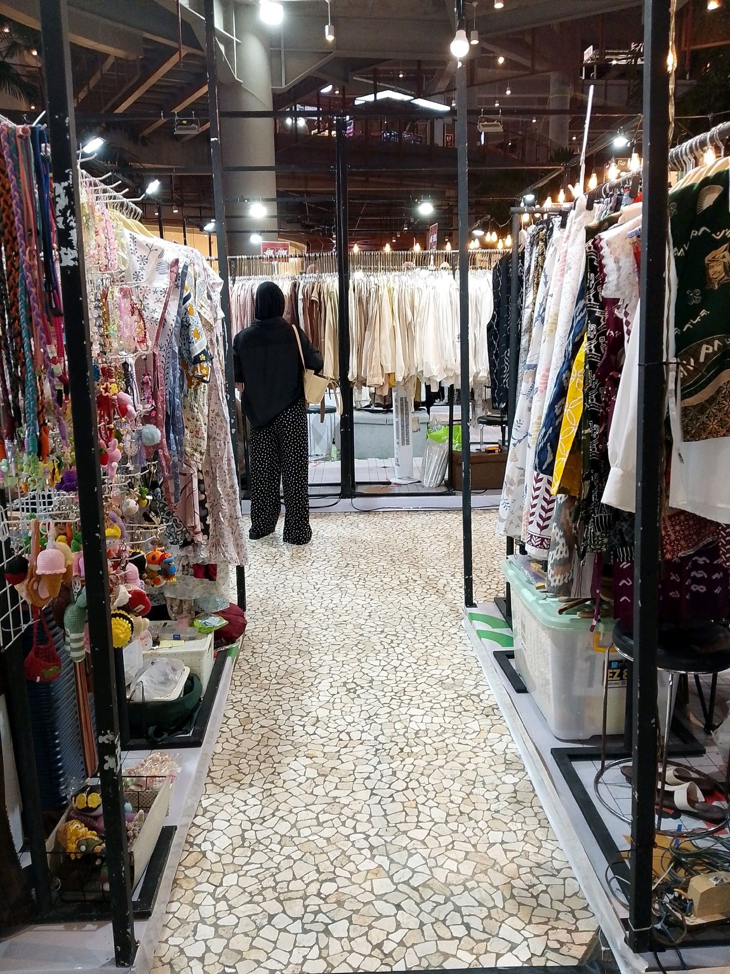 In a brightly lit indoor market stall in Makassar, Indonesia, a person wearing a black hijab, black shirt, and black and white polka dot pants stands facing away from the camera. They are looking at rows of clothing displayed on racks. The stall on the left is filled with colorful accessories, including necklaces, keychains, and knitted items. The stall on the right features a variety of patterned garments hanging on hangers. The floor is covered in a mosaic of small, irregular white tiles. The background shows the ceiling structure of the market, with exposed beams and numerous light fixtures.