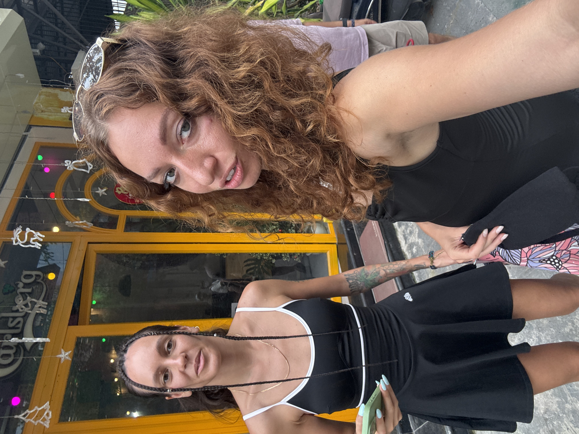 Two women are standing outside a building with a yellow facade, likely a restaurant or bar. The woman on the left has dark hair styled in braids and wears a black dress with white trim and a short, flared skirt. She holds a green smartphone in her left hand. She has a tattoo on her left arm. The woman on the right has curly, reddish-brown hair and is wearing a black tank top. Sunglasses are perched on her head. A blurred figure of a person wearing a light purple shirt and khaki shorts is visible in the background. Decorative lights, including Christmas-themed ornaments and circular colored lights, are hanging from the building. The overall setting suggests an outdoor urban environment.