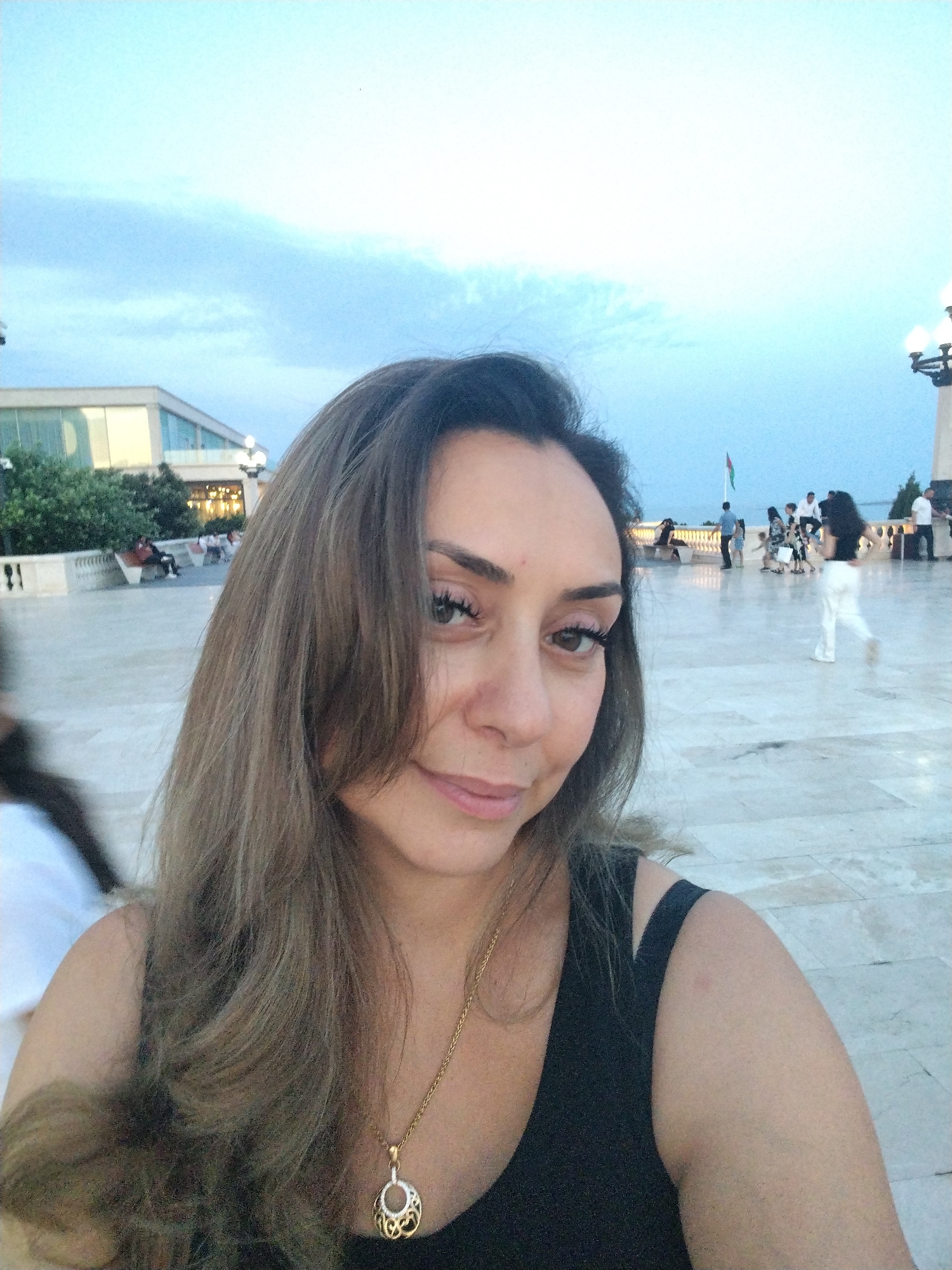 A selfie is taken outdoors in a paved plaza with a cloudy sky above. In the foreground, a woman with long, brown, layered hair is smiling. She is wearing a black tank top and a gold necklace with a circular pendant. In the background, several people are visible. To the left, a building with large windows is seen. Further back, a flag pole with a flag is situated near a railing. Other people are walking and sitting in the plaza, with streetlights visible in the distance. The scene appears to be in Sabail Raion, Azerbaijan.