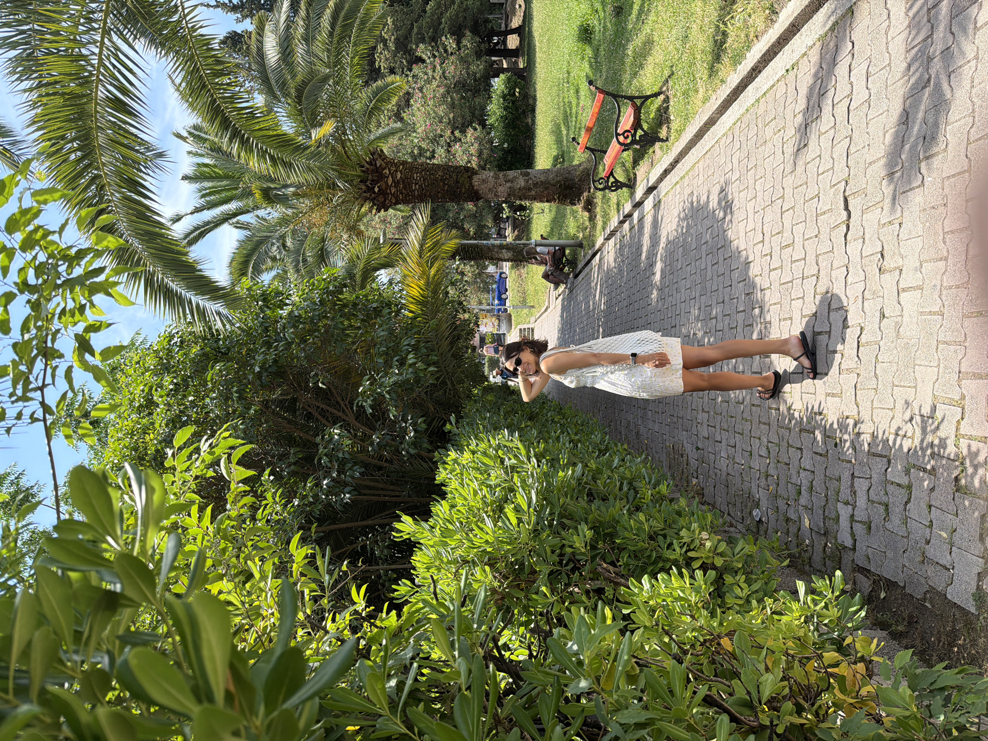 A woman in a white dress and black sandals stands on a paved path in a park, looking over her shoulder towards the camera. She has sunglasses on and her hand is raised to her hair. The path is lined with a hedge on the left and palm trees and grass on the right. A red bench is visible in the background. The scene appears to be in Budva, Montenegro, a coastal town known for its parks and palm trees.