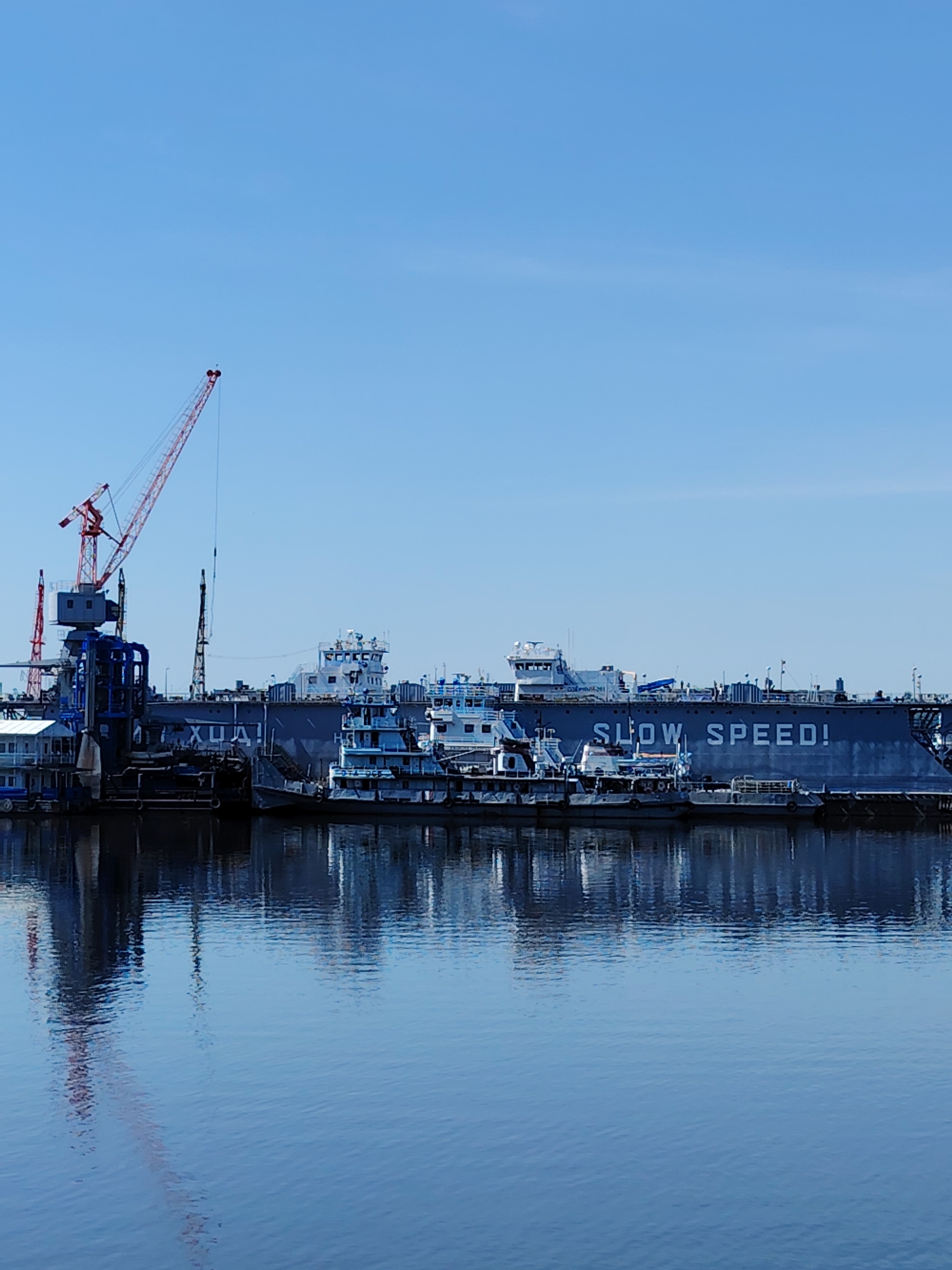 This image captures a waterfront industrial scene in Chaikovsky, Russia. The foreground is dominated by calm water reflecting the docked vessels and structures. Multiple tugboats and other watercraft are moored along a dark gray dock or barge, which has the phrase "SLOW SPEED!" painted in large white letters. A large floating drydock is also visible behind the tugboats. A red and gray crane is positioned on the left side of the image, towering over a blue structure with pipes. The sky is clear and blue.