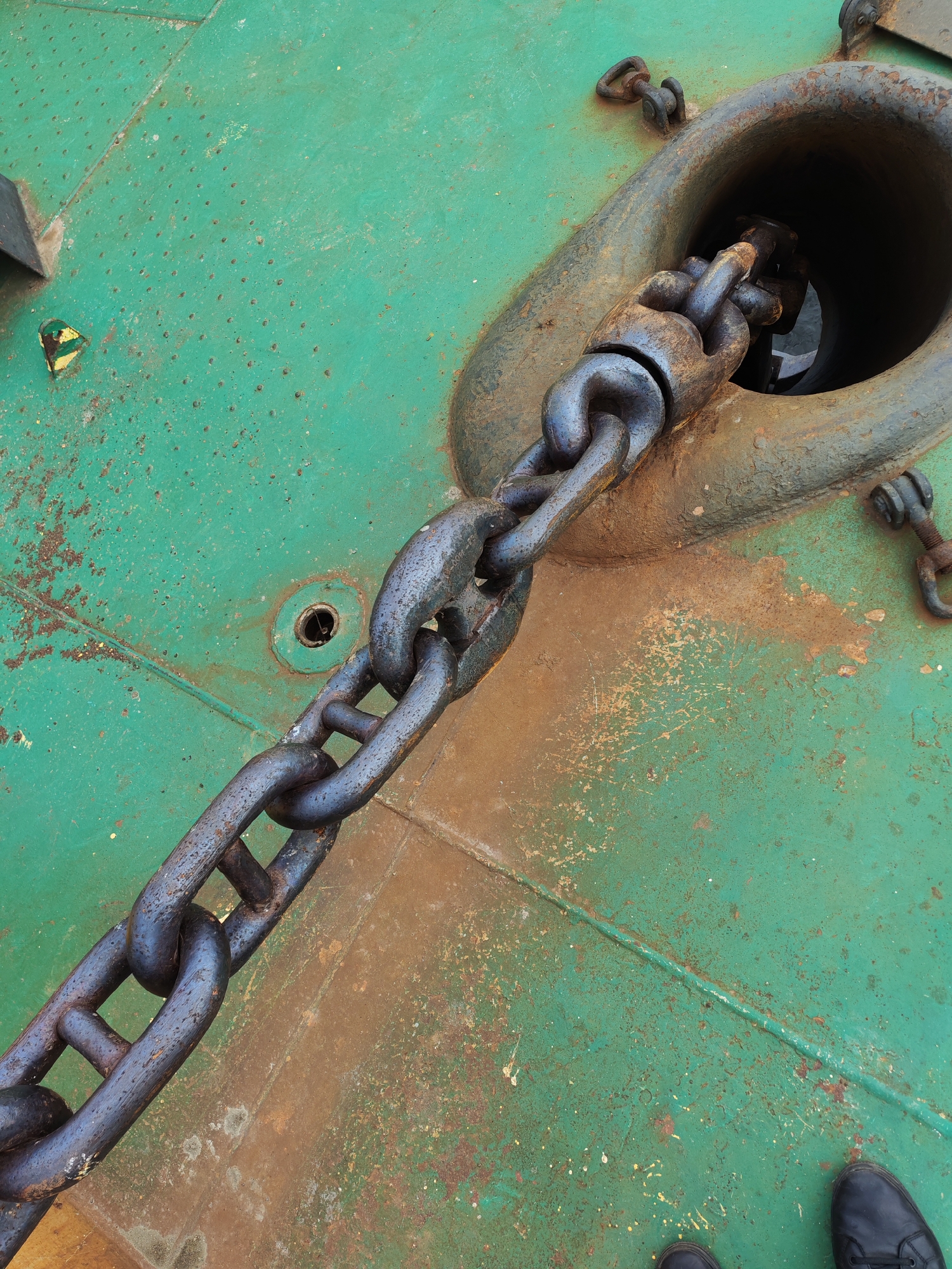 A close-up shot depicts a heavy, dark metal chain partially running through a large, circular hole reinforced with a dark, corroded metal ring. The chain extends diagonally across a green, textured surface showing significant wear and oxidation, with patches of brown rust. A smaller hole with a dark interior is also visible on the green surface near the large ring. At the bottom of the frame, a glimpse of two black shoes suggests a person is nearby. The overall scene presents an industrial or nautical context, suggestive of a ship or dock environment. There is no indication this photo was taken in Rybinsk, Russia.