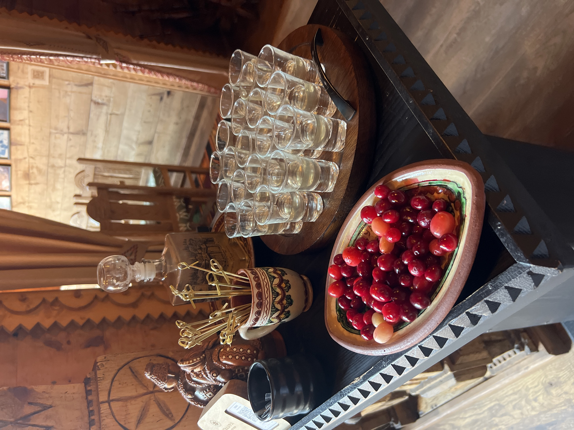 The image depicts a close-up of a rustic wooden table laden with a selection of beverages and snacks, likely in a traditional Ukrainian setting in Yaremche. In the foreground, a small, decorative ceramic bowl overflows with fresh red berries, possibly cranberries or lingonberries, interspersed with a few lighter-colored fruits. Beside the bowl, a dark ceramic vessel holds a bundle of thin wooden skewers. Further back, a wooden tray is neatly arranged with multiple small, clear shot glasses, each containing a light-colored liquid, presumably a spirit. To the left of the shot glasses, a decorative glass decanter with an ornate stopper stands on the table, filled with a similar liquid. The background reveals wooden paneled walls and a glimpse of traditional wooden furniture, including a chair and curtains, contributing to the overall rustic ambiance. The lighting suggests an indoor setting, possibly a restaurant or guesthouse.
