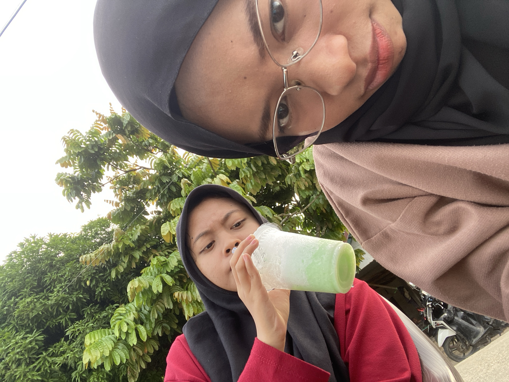 This is a selfie-style, eye-level, wide-angle shot featuring two young women in a tropical outdoor setting. The woman in the foreground, on the left, wears a black hijab and a red long-sleeved shirt. She is holding a translucent plastic cup filled with a pale green, frothy beverage and a straw. She is looking down at the drink as she sips it.
Behind her and slightly to the right, partially obscuring the background, is the second woman. She is wearing a black hijab and a light brown cardigan or jacket over a darker garment. She has glasses and is looking into the camera. Her face is positioned close to the lens, giving a sense of intimacy and immediacy to the photograph. Her lips are slightly parted and appear to have light pink lipstick.
The background consists of lush green foliage from trees, suggesting a warm climate. A portion of a motorbike is visible in the lower right quadrant, along with what appears to be a paved surface, possibly a road or parking area. The lighting indicates it is daytime, likely late morning or afternoon, with overcast skies, as there are no strong shadows. The overall mood is casual and relaxed, possibly depicting friends enjoying a drink outdoors in Indonesia. There is no visible text in the image.