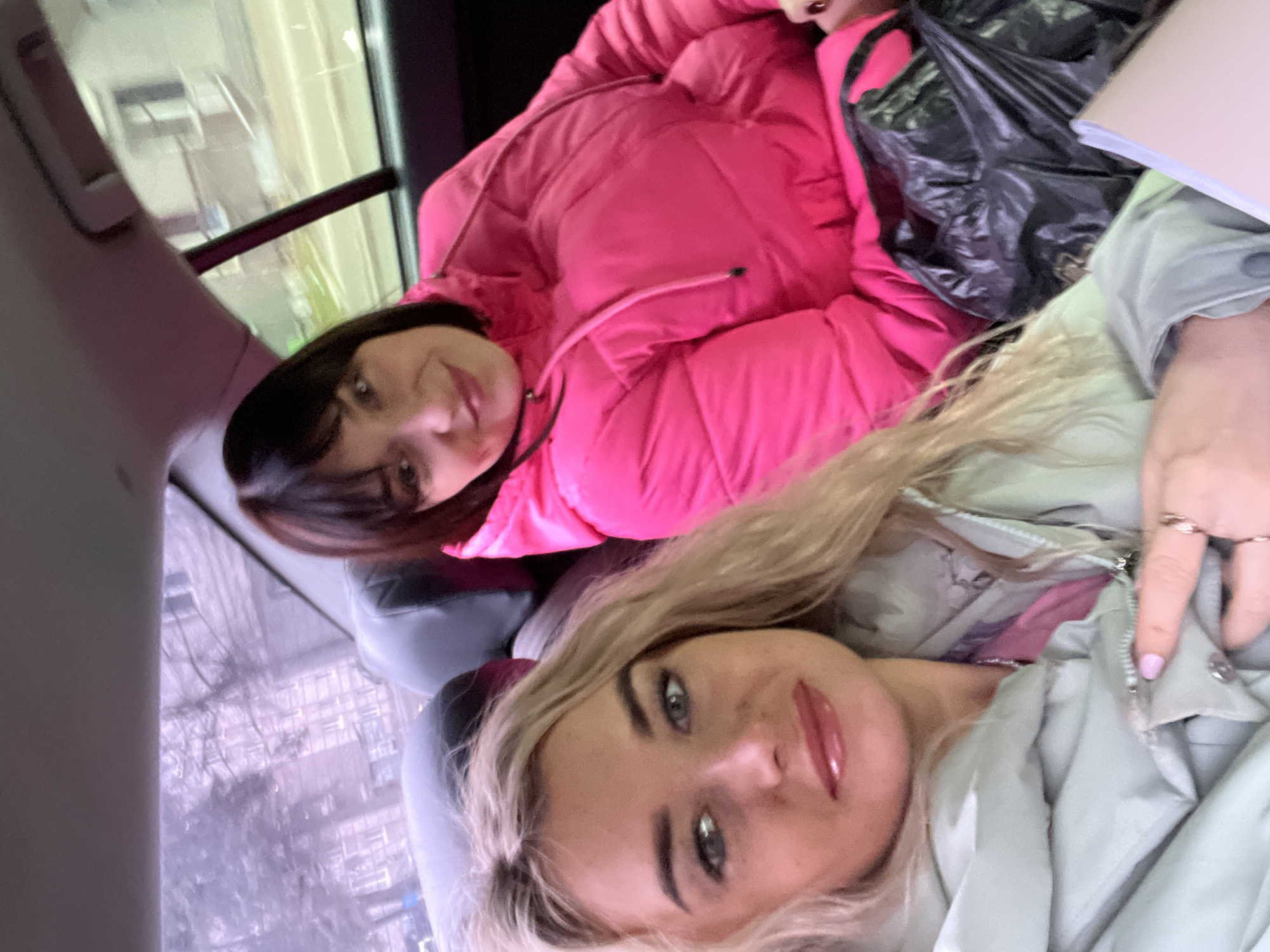 Two women are posing for a selfie. One has blonde hair and is wearing a light green jacket. The other has dark hair with bangs and is wearing a pink puffer jacket. They appear to be inside a vehicle, possibly a car or a bus, as suggested by the window behind them. The location is Izhevsk, Russia, based on the view through the window, which shows buildings and trees typical of a Russian urban landscape. One woman’s hand is visible near the zipper of her jacket. A black plastic bag is next to the woman in the pink jacket.