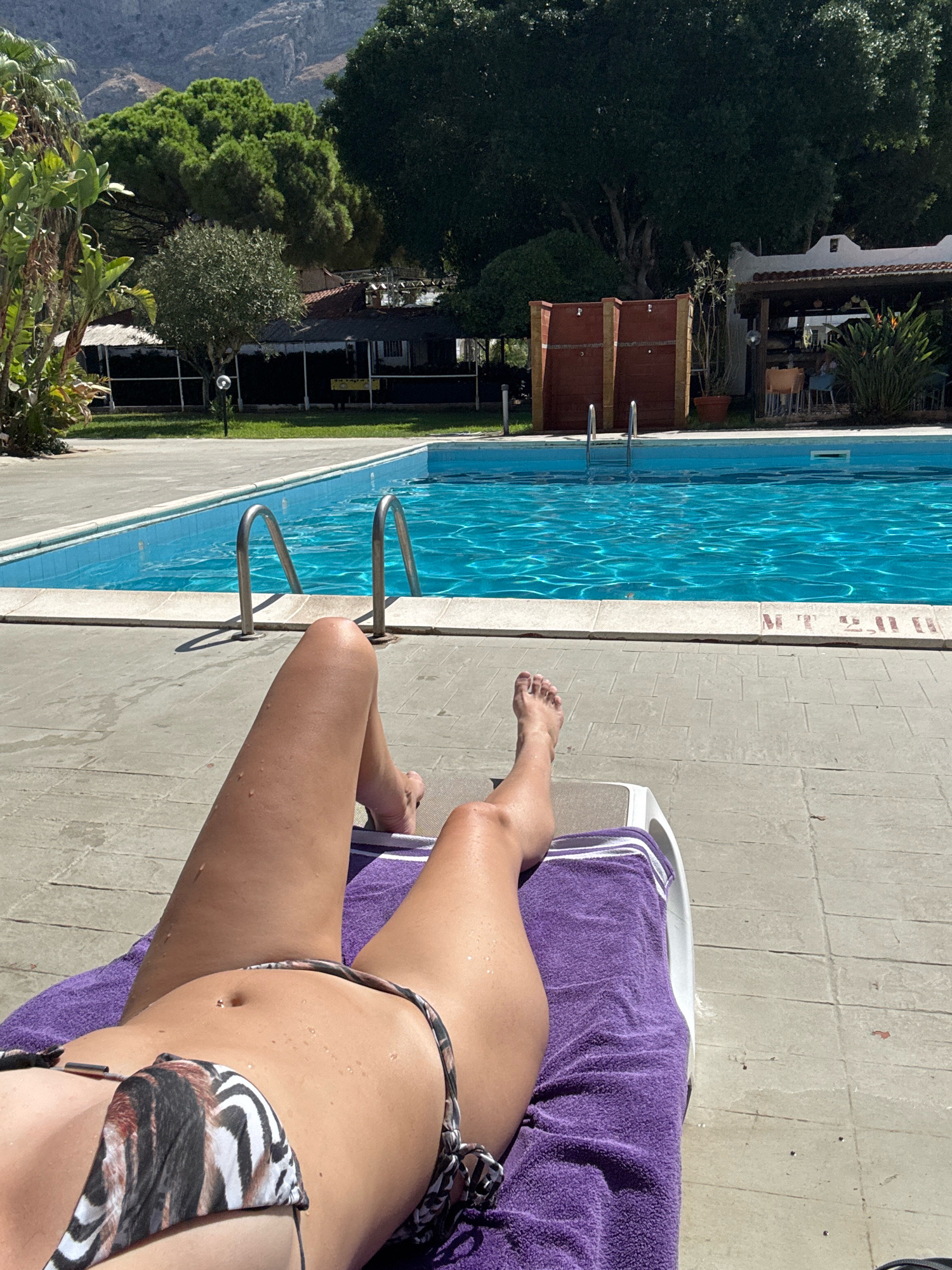 A woman lies on a lounge chair by a swimming pool, her legs and torso visible. She is wearing a bikini with a zebra-like pattern and a belly button piercing. A purple towel is draped over the lounge chair. The swimming pool is filled with bright blue water and has metal railings. In the background, there are trees, buildings, and a mountainous landscape. The scene appears to be outdoors in a sunny location. This is likely a hotel or resort area in Isola delle Femmine, Italy, given the Mediterranean vegetation and architecture.