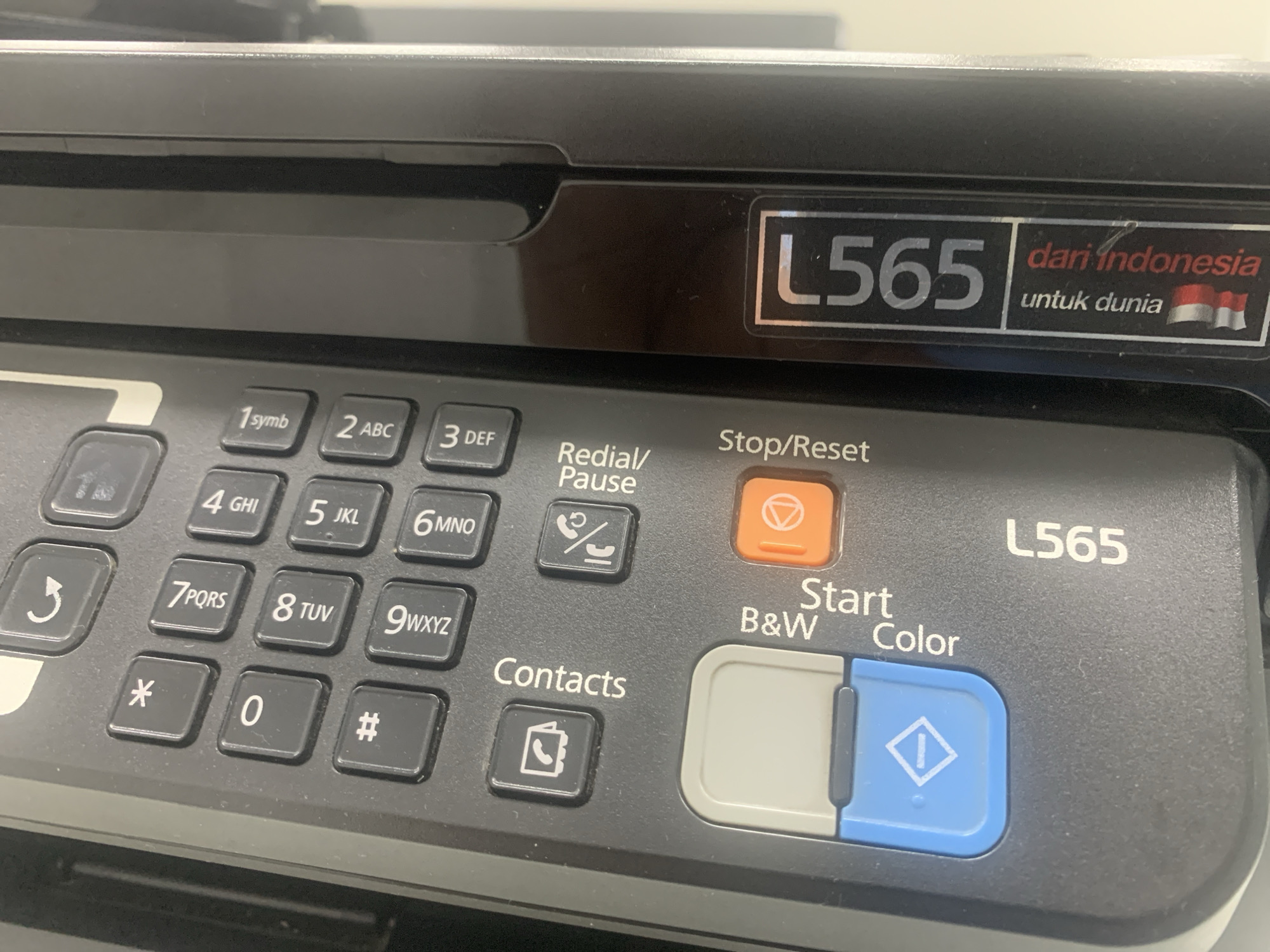 This is a close-up, eye-level shot of the control panel of a black printer, likely an Epson L565 model, based on the visible branding. The printer is positioned on a flat, light-colored surface, possibly a desk, in an indoor setting. The control panel features a numerical keypad with buttons labeled 0 through 9, as well as asterisk (*) and pound (#) symbols. Adjacent to the keypad are function buttons, including "Redial/Pause," "Stop/Reset," "Contacts," and two large, distinct buttons labeled "Start B&W" and "Color," with an icon of a square with a vertical line inside on the color button. To the right of the numerical keypad and above the start buttons, the model number "L565" is printed in silver. To the upper right of the panel, a silver rectangular label displays "L565" and to its right, a red and white Indonesian flag with text in red and black reads "dari indonesia untuk dunia." The lighting is artificial and appears to be from overhead, creating a slightly warm tone. There are no people or animals visible in the image, nor any clear indication of the city of Palembang, Indonesia, beyond the text on the label.
