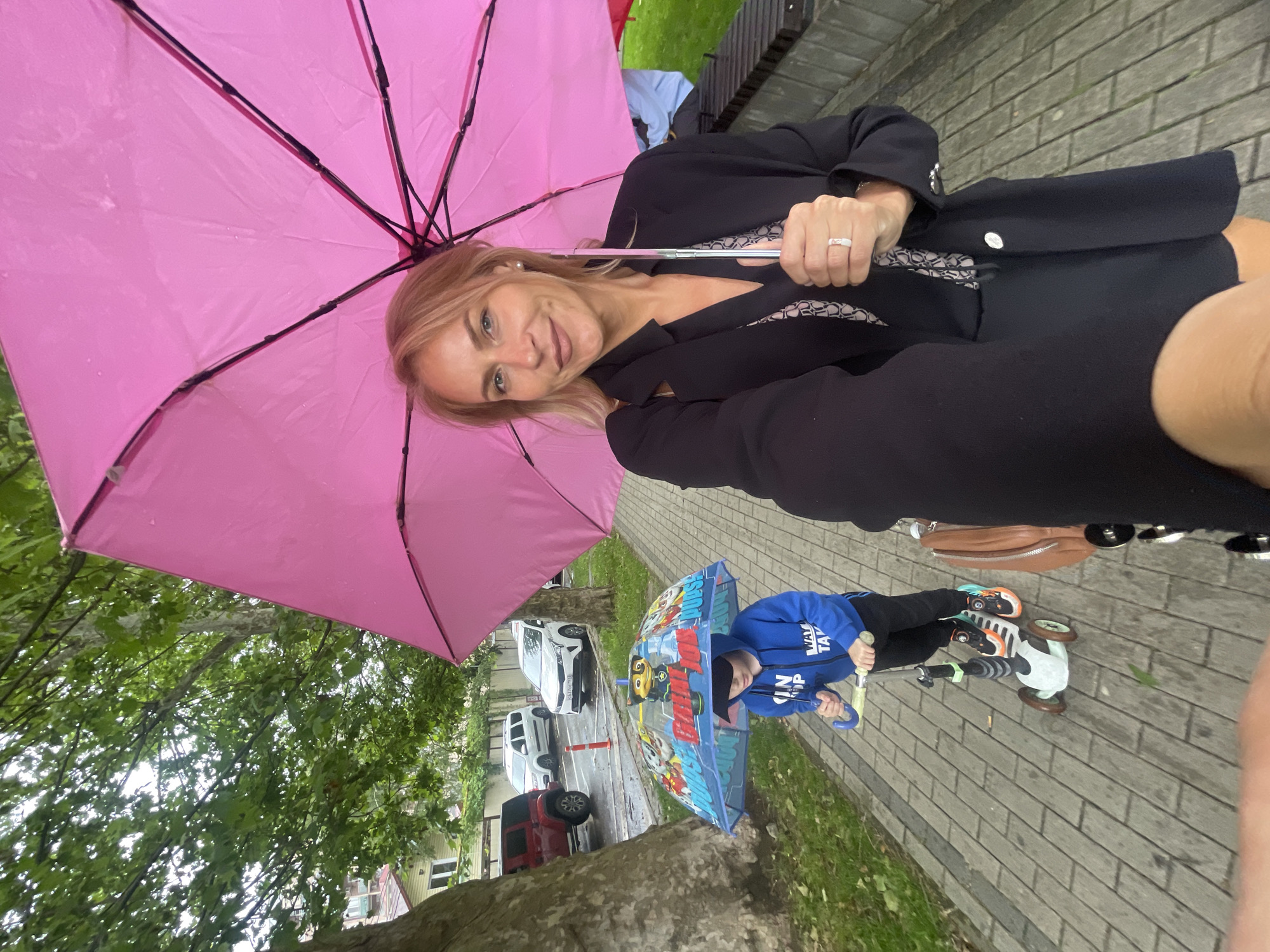 A woman with blonde hair and a black blazer stands under a pink umbrella on a brick pathway. A child in a blue hoodie rides a scooter under a Paw Patrol-themed umbrella. Cars are parked along the street, suggesting a residential area. The location is Sochi, Russia, as indicated by the license plate "777RUS" visible on one of the parked cars.