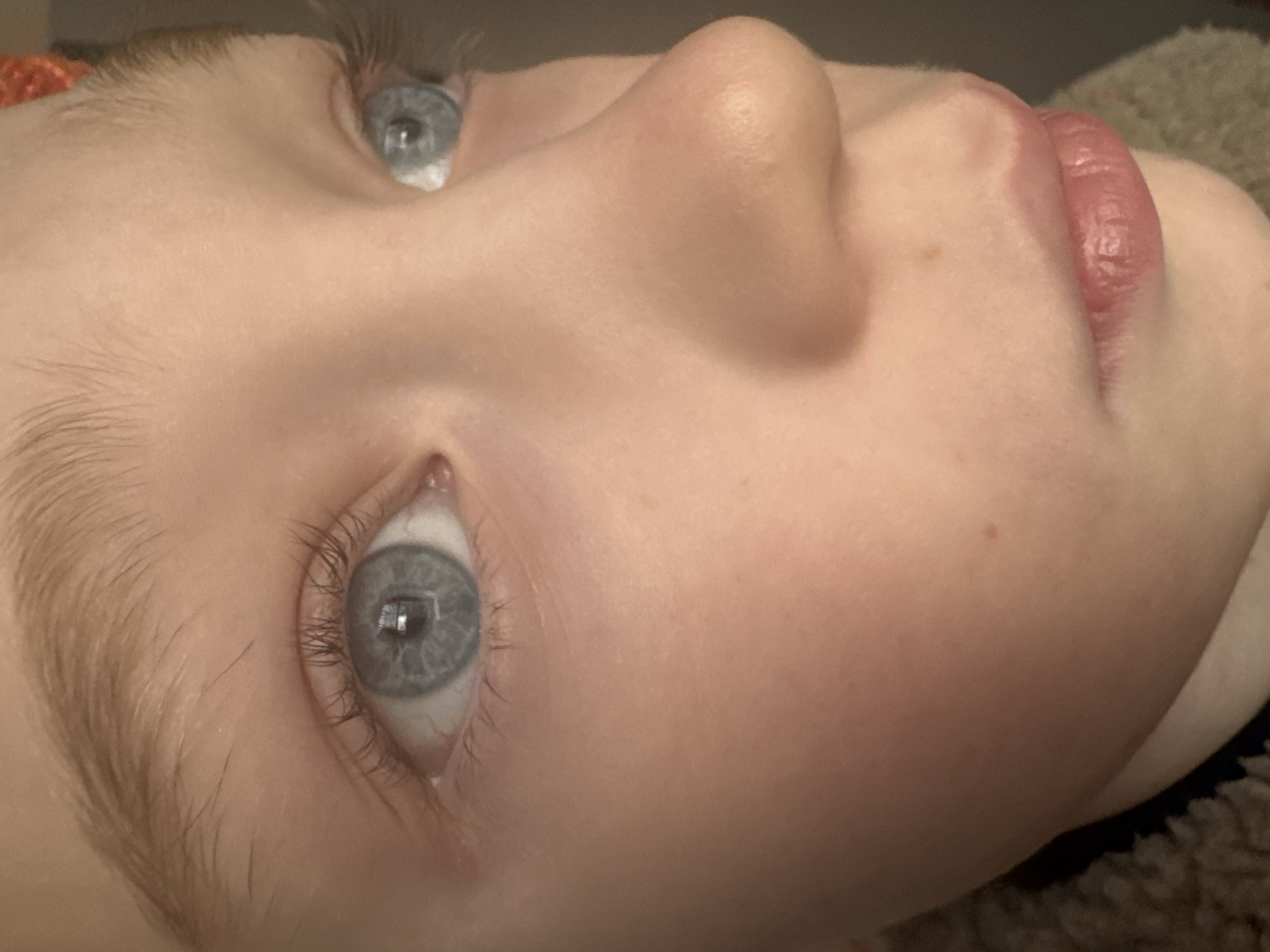 The image presents a close-up, eye-level portrait of a single individual, likely a child or young adolescent, focusing on their facial features.
**Location:** The specific location in Votkinsk, Russia, cannot be determined from the visual information provided. The background is indistinct and out of focus, suggesting an indoor setting.
**People:** One person is depicted, presenting fair skin with some minor redness on the cheeks and nose. Several small, dark freckles or moles are visible on the left cheek and above the upper lip. The individual has light blue eyes, with distinct dark pupils. Reflections of a rectangular light source, possibly a window or screen, are present in the irises of both eyes. Eyelashes are visible on both upper and lower lids, and the eyebrows are light brown or blonde. Light brown or blonde hair is also visible near the right temple. The nose is prominent, and the lips are naturally pink and appear slightly parted or closed, showing some natural creases.
**Subjects/Objects:** The primary subject is the face of the individual. A textured, fuzzy garment, possibly fleece or sherpa material, in a light brown or beige color is visible on the lower left and lower right edges of the frame, consistent with clothing. A small, blurry portion of an orange or reddish object, potentially a hair accessory or part of clothing, is visible near the upper right corner of the frame.
**Actions:** The individual is looking forward, directly or slightly to the right of the viewer's perspective. No other actions are discernible.
**Interactions:** No interactions are observable as only one individual's face is present within the frame.
**Environment:** The background is entirely out of focus and appears dark and uniform, offering no specific details about the surrounding environment other than indicating an interior setting with a consistent, soft light source.