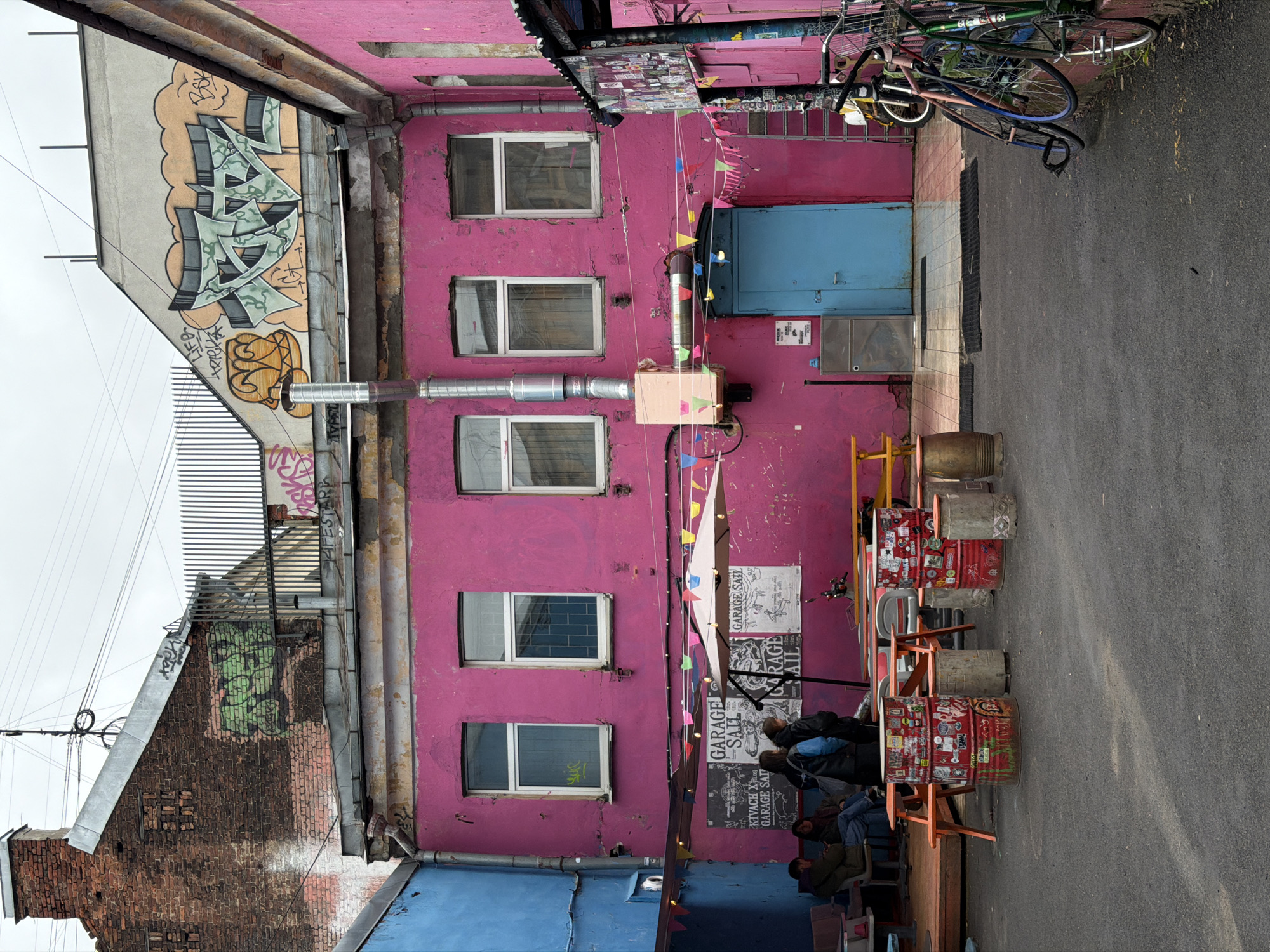 The image depicts a narrow courtyard or alleyway in Saint Petersburg, Russia. Dominating the scene is a tall, narrow building painted a vibrant pink. The building has five windows arranged vertically. A blue door is visible on the upper right, along with what appears to be a metal box. A metal ventilation system runs down the building's exterior. Colorful bunting is strung across the courtyard.
On the left side of the image is another building with visible graffiti. It is multi-storied, showing brick and plaster construction.
In the foreground, there is a makeshift seating area with metal barrels repurposed as tables and wooden benches. Several people are seated, suggesting a casual meeting or social space. Stickers cover the barrels. Bicycles are leaning against the pink building in the background. The ground is paved asphalt.