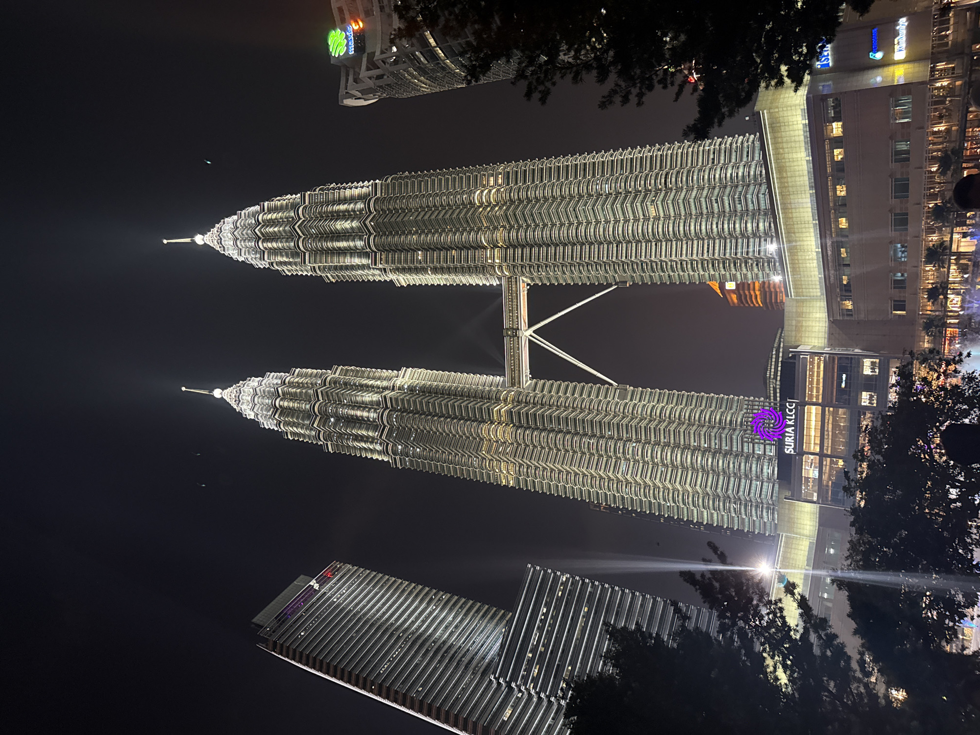 This night photograph captures the iconic Petronas Twin Towers in Kuala Lumpur, Malaysia, illuminated against a dark sky. The two towering structures, seen from a low angle, stand prominently, showcasing their intricate, modern architecture and distinctive spires. Both towers are extensively lit with white and yellowish lights, highlighting the detailed patterns of their facades. The skybridge connecting the two towers at a mid-level is also clearly visible and brightly lit.
In the foreground and scattered around the base of the towers are dark silhouettes of trees and foliage, adding depth to the scene. Several other modern buildings are present; one to the right, adjacent to the towers, features numerous illuminated windows and the prominent text "SURIA KLCC" with a purple spiral logo, indicating the shopping mall at the base. Further right, text like "isetan" and "community" is discernible on another building. In the bottom left, a smaller, illuminated skyscraper is visible, featuring a green logo and possibly the text "Maybank". Another building in the distance also shows a green "Maybank" logo and "SSTi" text.
The time of day is clearly night, with the artificial lights creating a striking contrast against the almost black sky. There are faint atmospheric glows around the bright lights. No people are visible, and no specific activity or event is depicted beyond the static display of the illuminated architecture. The overall impression is one of modern urban grandeur and a clear evening with no adverse weather conditions like rain or heavy fog.