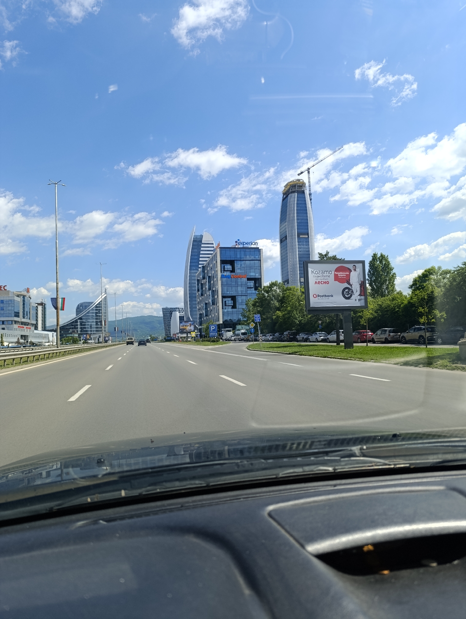 Captured from within a vehicle, the image portrays a scene on a highway leading towards the city of Sofia, Bulgaria. Modern high-rise buildings dominate the background, including the Capital Fort complex and the Sofia Airport Center. The cityscape is accented by a towering skyscraper under construction. The highway stretches into the distance, with occasional cars visible in the lanes. The sky is blue with scattered clouds, indicative of a sunny day. On the right side of the road, a billboard displaying an advertisement for Postbank is noticeable. The foreground of the image is dominated by the car's dashboard and windshield, suggesting the photo was taken while driving.