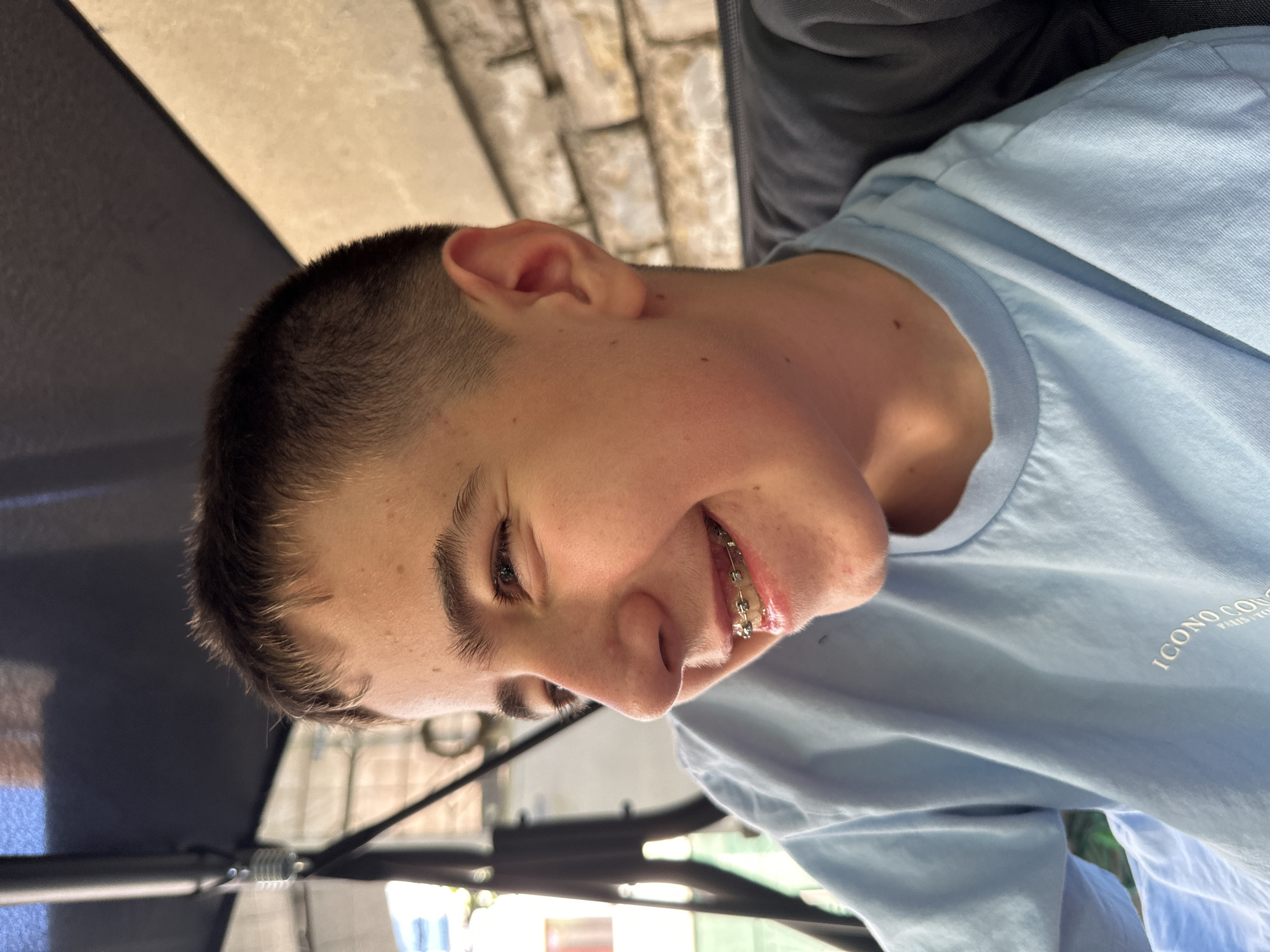 A fair-skinned young man is smiling, showing dental braces. He is wearing a light blue t-shirt with the faded text "ICONO CONCEPT PARIS TOKYO" on the right breast. The subject has a short, dark haircut. The picture appears to be taken outdoors, under the cover of an umbrella. A building facade made of light-colored brick is visible in the background. The location of this photo is Unknown, Unknown.