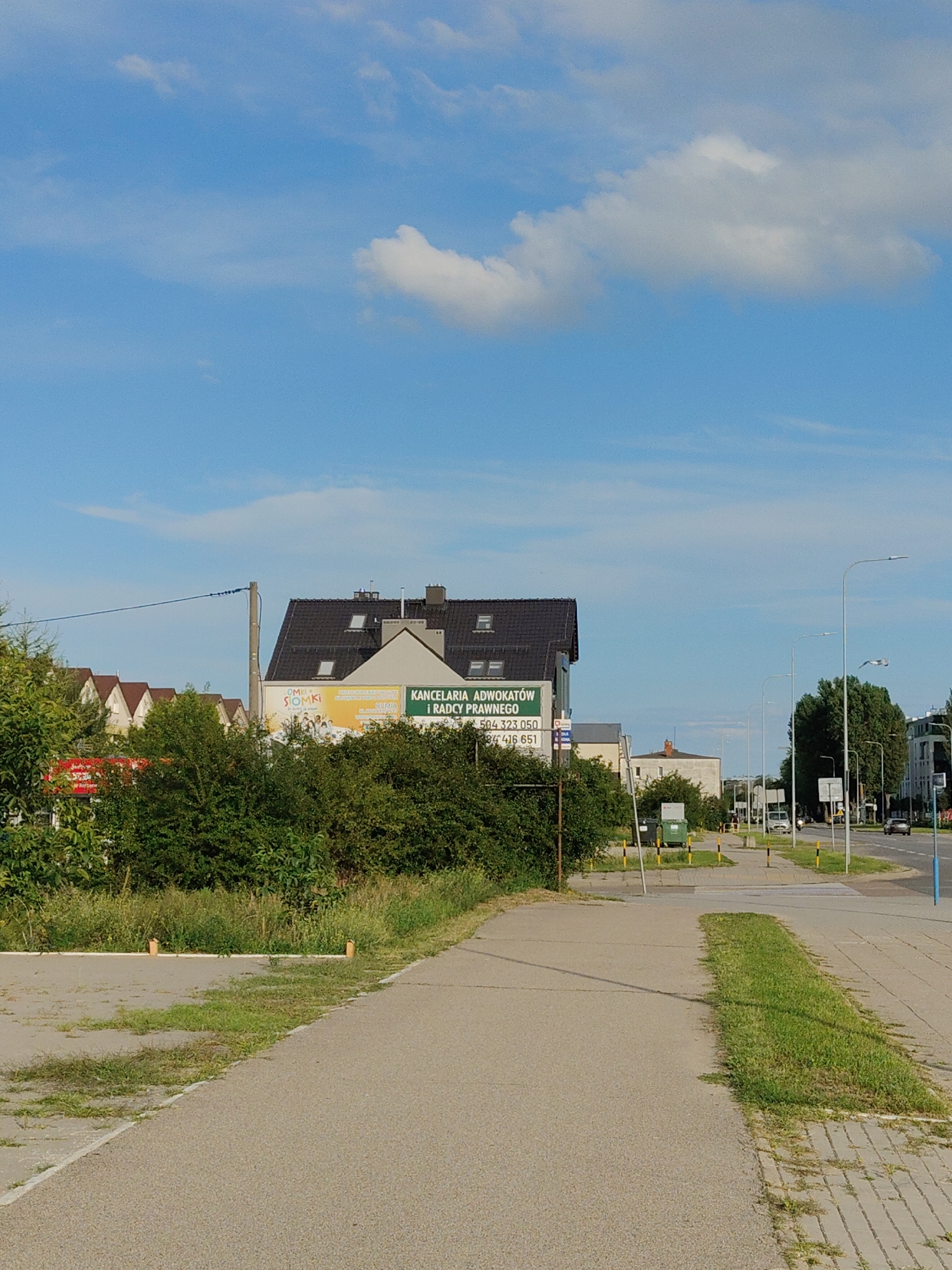 The image shows a street scene in Rumia, Poland, on a sunny day with scattered clouds. In the foreground, a paved pedestrian path runs parallel to a road. To the left of the path is a grassy area with some trees and bushes, and further left, a row of houses with terracotta roofs. In the middle ground, a building with a dark pitched roof displays a green sign that reads "KANCELARIA ADWOKATOW / PRAWNEGO" (Lawyer's Office) along with contact information. Adjacent to this building, a billboard with the text "SIOMKI" is visible. The road is lined with streetlights and some vehicles, including a car, are visible in the distance. The overall atmosphere is that of a typical urban or suburban street.