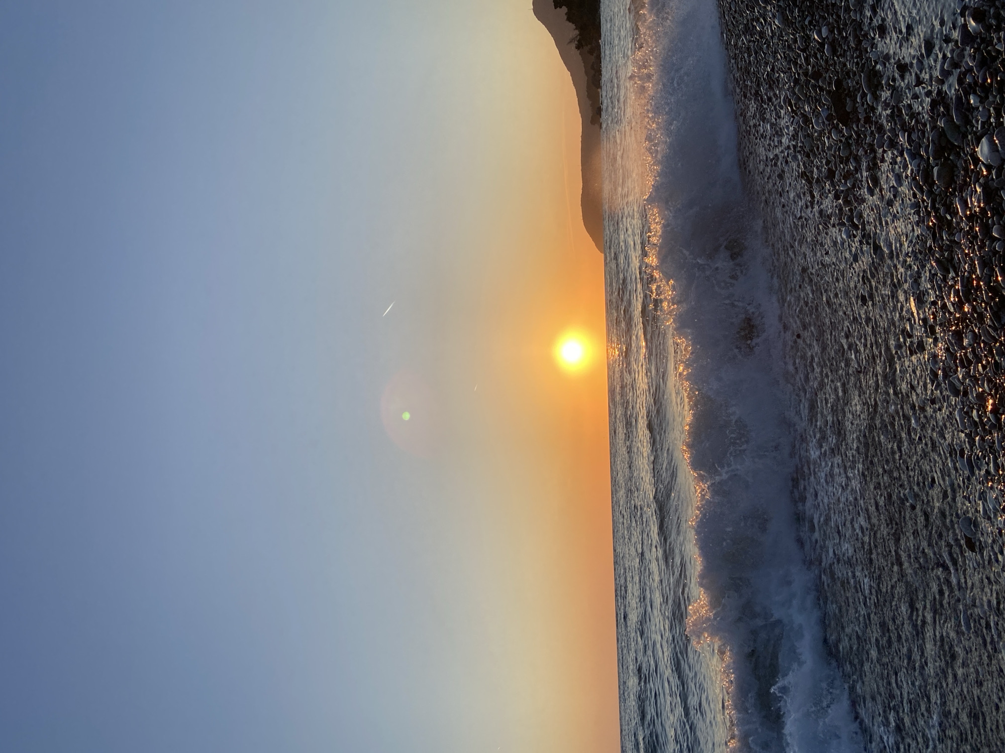 The image captures a picturesque coastal scene at either sunrise or sunset in Bar, Montenegro. The sun, a vivid yellow-orange orb, is positioned low on the horizon, casting a warm, golden light across the entire frame. The sky transitions from a soft, clear blue at the top left to warm hues of orange and pink around the sun. A subtle airplane contrail is visible in the upper left quadrant of the sky, accompanied by a distinct green lens flare to the left of the sun.
To the right, the sea meets a dark pebble beach. Waves are breaking, their foamy crests and the water's surface shimmering with intense golden reflections from the low sun. The beach itself is composed of numerous dark, smooth pebbles, many of which are wet and reflect the warm light, creating intricate patterns of highlights. A dark, silhouetted mountain range or large hill rises in the background along the coastline, partially obscuring the horizon to the right of the sun. The scene evokes a sense of tranquility and natural beauty. There are no people or visible text in the image.