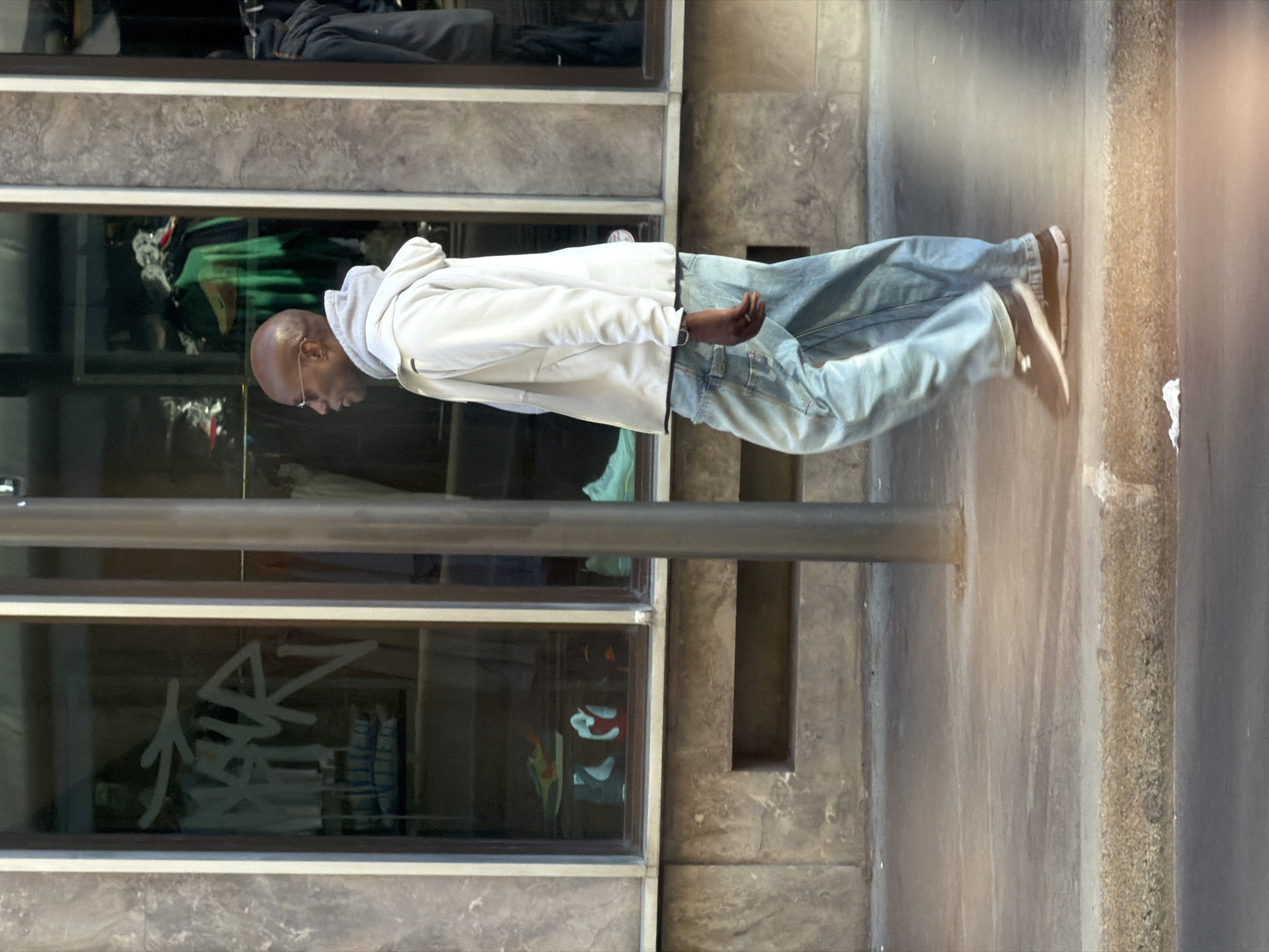 An outdoor, eye-level shot, originally oriented vertically but depicting a horizontal scene, captures a man walking or standing on a city sidewalk in what is identified as Paris, France. The man, who is bald and wears glasses, is dressed in a light-colored, possibly white or light grey, hooded jacket, light blue jeans, and dark sneakers with white soles. A wristwatch is visible on his left wrist. He is positioned next to a dark, metallic pole that extends from the bottom to the middle of the frame.
To the man's left is a building facade with several large windows, likely belonging to a shop or commercial establishment. These windows display reflections of the surroundings and vague forms of merchandise, possibly clothing, within. On one of the lower windows, white graffiti or stylized writing, which appears to include characters resembling "14Z" or similar abstract shapes, is visible.
The ground is a paved surface, likely concrete or stone, suggesting an urban street or pedestrian area. Bright daylight illuminates the scene, with a notable light flare or strong highlight on the far right, indicating direct sunlight. The overall setting is an everyday urban environment, with the architectural elements of the building and sidewalk consistent with a Parisian streetscape. The man's expression is not clearly discernible as he is looking downwards or to his side.