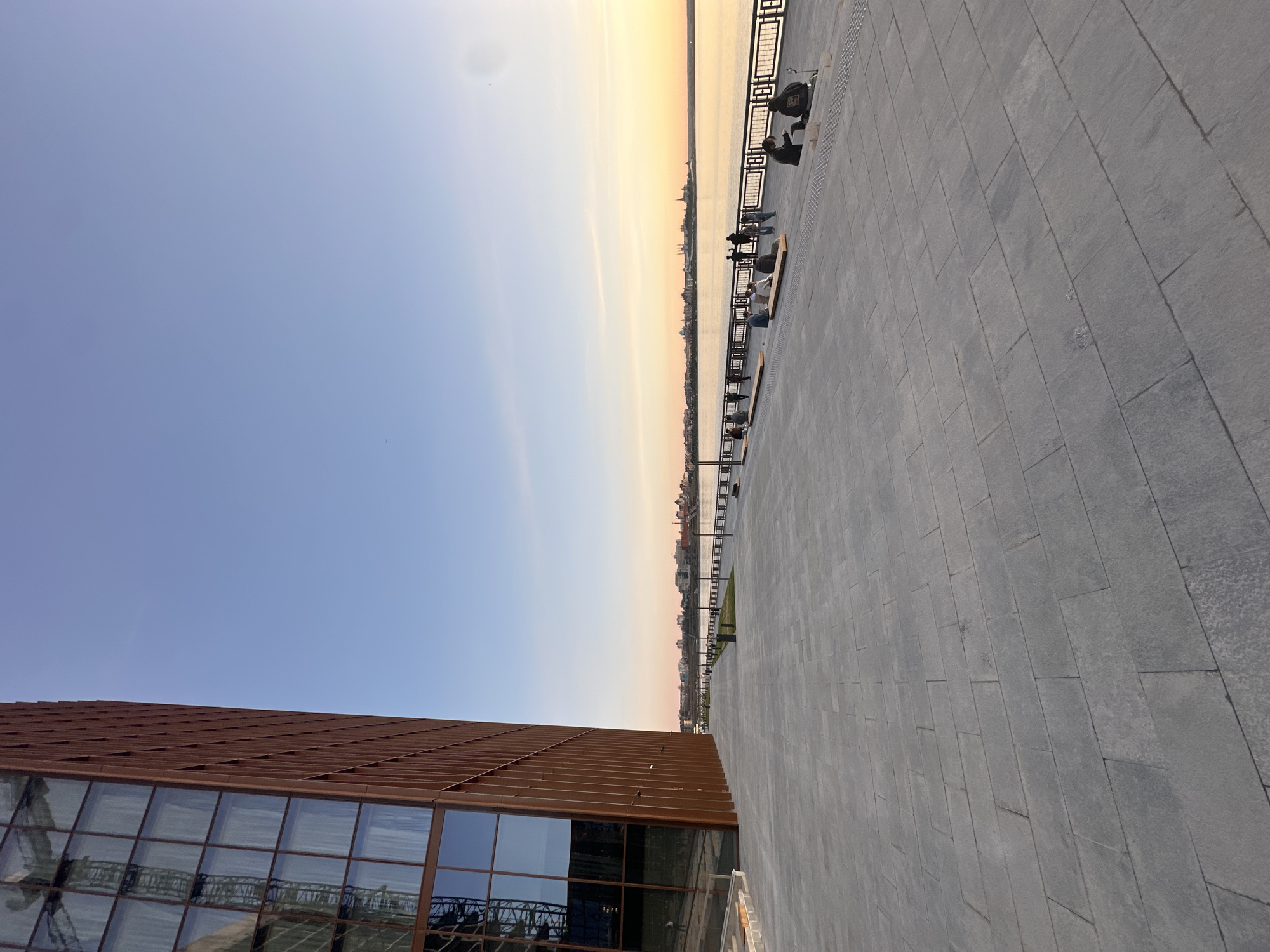 This image captures a wide, modern promenade or embankment in Kazan, Russia, during what appears to be either sunset or sunrise. The sky displays a clear gradient, transitioning from a soft blue at the top to a warm orange and yellow glow at the horizon, where it meets a tranquil body of water.
The scene is dominated by a broad, grey paved walkway on the right, made of rectangular stone tiles. A dark metal railing with decorative elements runs along the edge of the promenade, separating it from the water. Several people are visible along this railing, some seated on benches or low structures, while others stand, seemingly observing the view. In the distance, across the calm water, the distinct silhouette of a city skyline, characteristic of Kazan, can be seen under the vibrant sky.
On the left, a modern building with a unique architectural design occupies a significant portion of the foreground. It features a textured, reddish-brown facade and large, reflective glass windows that mirror parts of the sky and the surrounding environment. The overall atmosphere is serene, with individuals enjoying a leisurely moment by the waterfront. No explicit text is visible in the image.