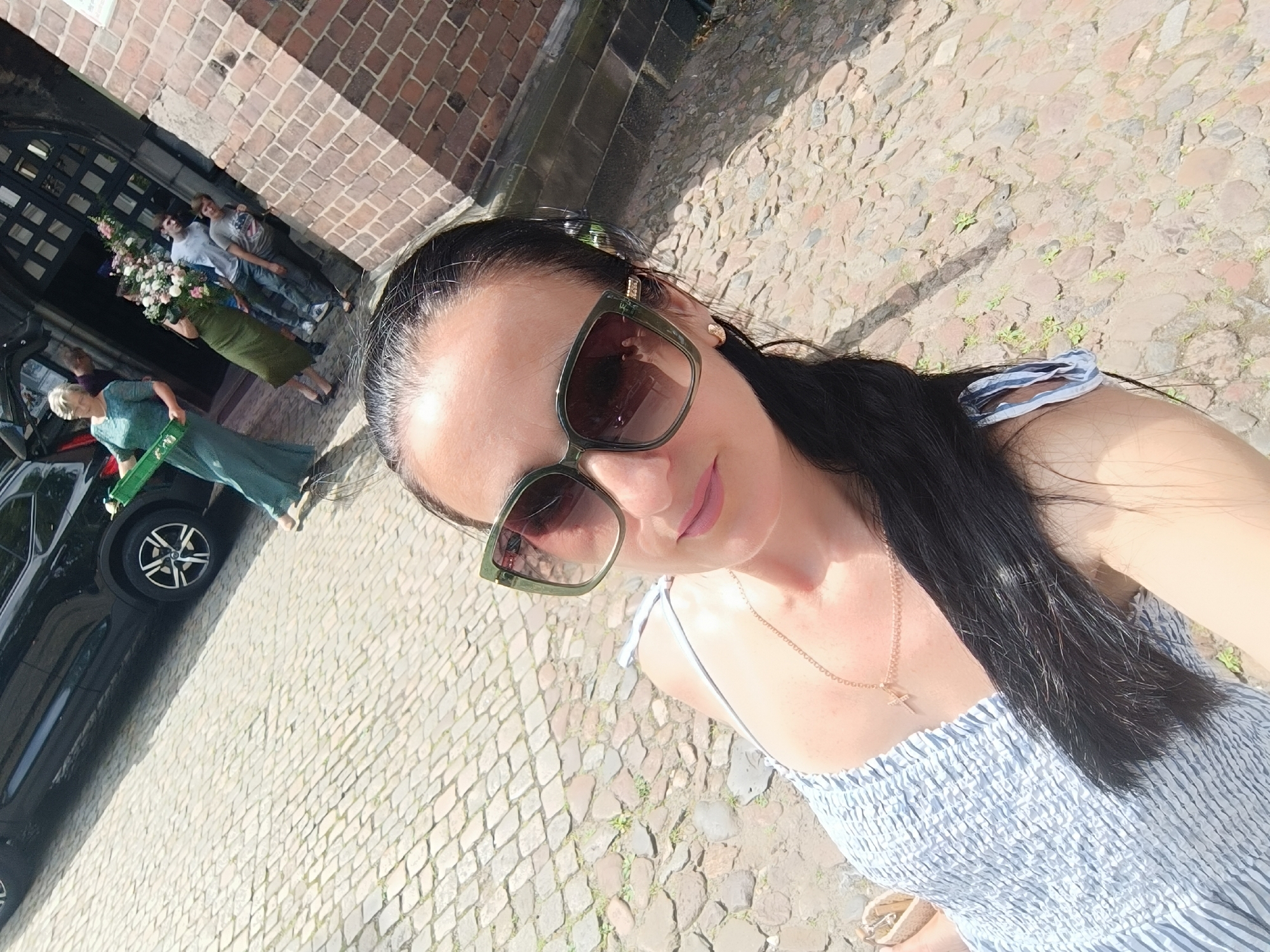 The image shows a woman taking a selfie outdoors. She is wearing sunglasses and a blue and white striped dress. In the background, several people are standing near a building with a brick facade and an arched doorway. One person is holding a large bouquet of flowers. A black car is parked to the left of the group. The ground is paved with cobblestones. The scene appears to be in a historic city square or near a church in Gliwice, Poland. The lighting suggests it is daytime, likely sunny. The woman in the foreground is the primary subject of the selfie, with the background offering context.