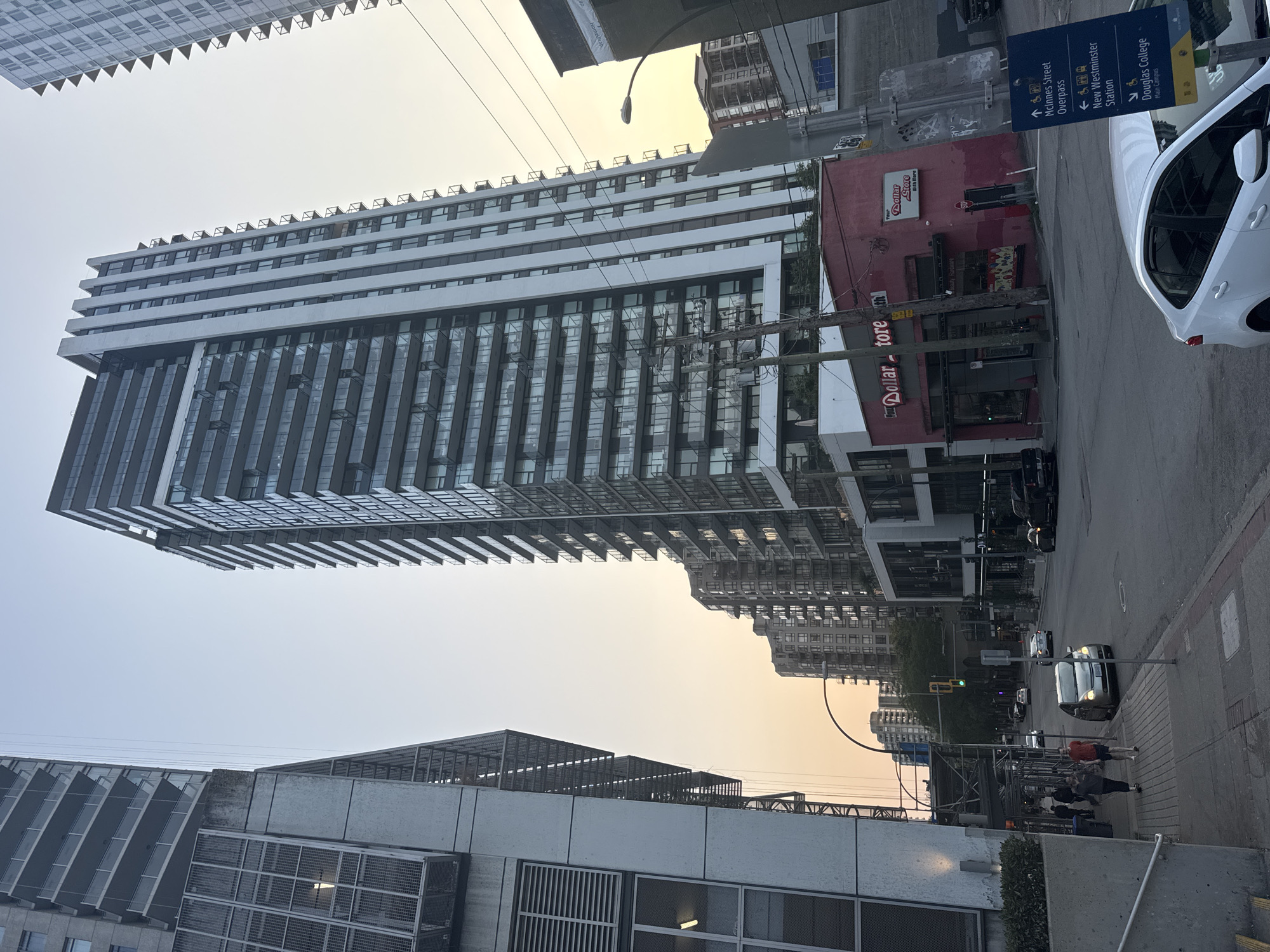 The image depicts a street scene in New Westminster, Canada, likely in the downtown area. The foreground is dominated by a sidewalk with a few pedestrians walking and a white sedan parked. To the left, a modern, grey building rises, featuring prominent ventilation grates and windows. A taller, glass-fronted residential building stands to the right of the grey structure, showcasing multiple balconies. Further into the background, other multi-story buildings line the street, with a prominent red building labeled "Your Dollar Store" visible. Overhead, power lines and streetlights are present. Traffic signals and street signs, including one indicating "McInnes Street Overpass" and "New Westminster Station," are also visible. The sky is a soft orange and blue, suggesting either sunrise or sunset. The overall impression is an urban environment during a transitional time of day.