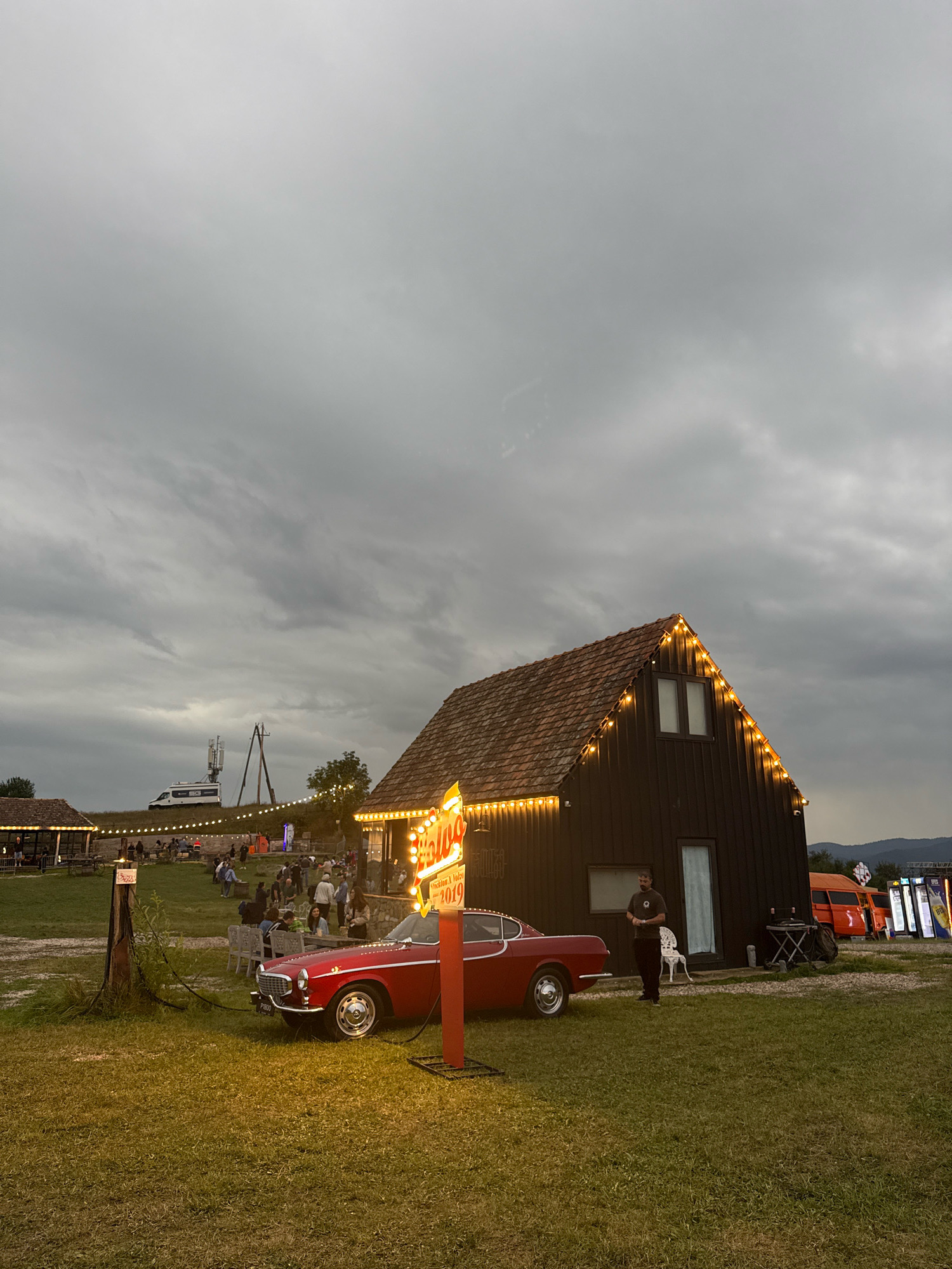 The image depicts an outdoor gathering or event in Orbeti, Georgia, under a cloudy sky. In the foreground, a vibrant red vintage car, identified as a Volvo P1800, is parked on a grassy area. A neon sign with the word "Haloo" and the year "2019" is illuminated and stands next to the car.
Behind the car, a dark wooden building with a tiled roof is visible. The building is decorated with string lights along its eaves. A person stands near the entrance of this building. Further back, several smaller wooden structures with covered seating areas are set up, with people gathered at tables and mingling. In the middle ground, a white van and a tall antenna tower are present. The background shows a hilly landscape with mountains in the distance. The overall atmosphere suggests a casual event or festival.