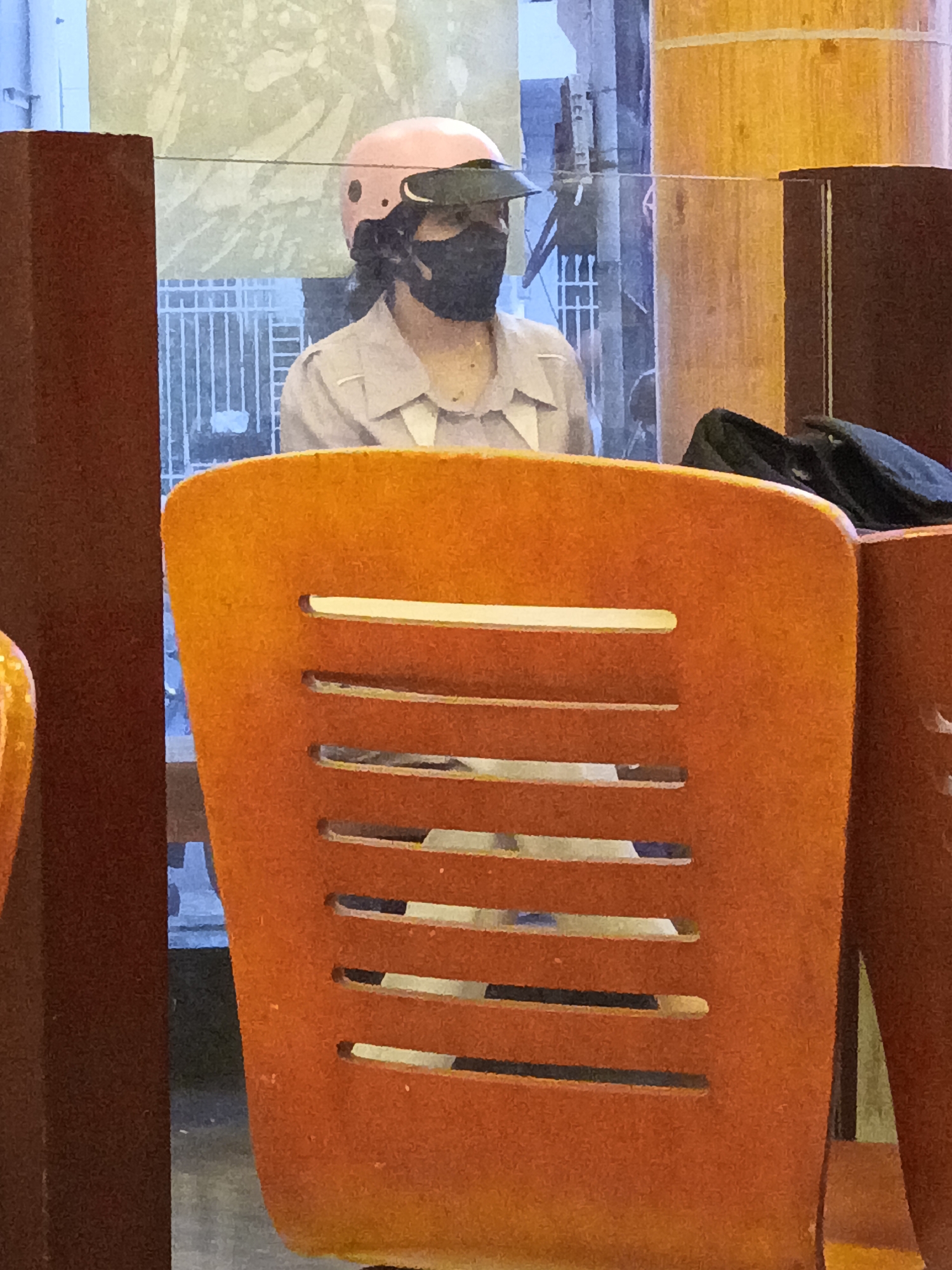 A person wearing a pink helmet and a black face mask is sitting behind an orange chair. The person appears to be wearing a light-colored collared shirt. The background shows a building with windows and some greenery. The scene appears to be outdoors, possibly in a cafe or restaurant with outdoor seating. The lighting suggests it might be daytime. There is no visible text in the image.