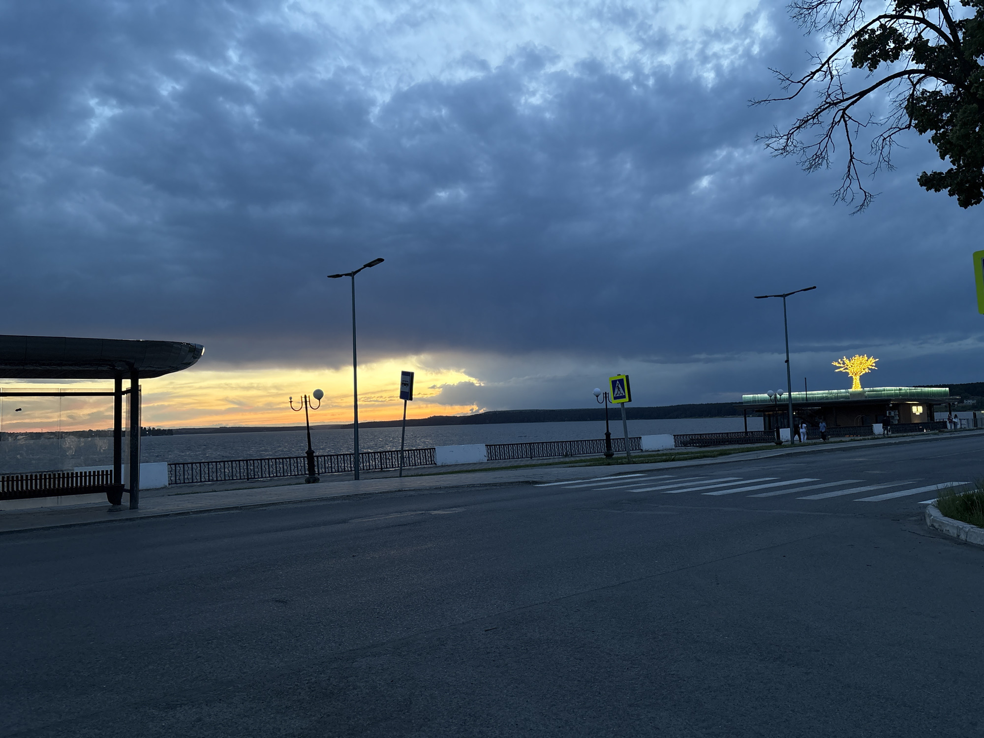 The image depicts a street scene in Votkinsk, Russia, at dusk or dawn. The foreground is dominated by a paved road with a pedestrian crossing. To the left, a bus stop provides shelter. A wrought-iron fence runs along the waterfront, separating the road from the water. Streetlights illuminate the area. In the background, the sky is overcast with dark clouds. A building or structure with a decorative tree illuminated with lights sits on the waterfront. Several people are present near the illuminated structure.