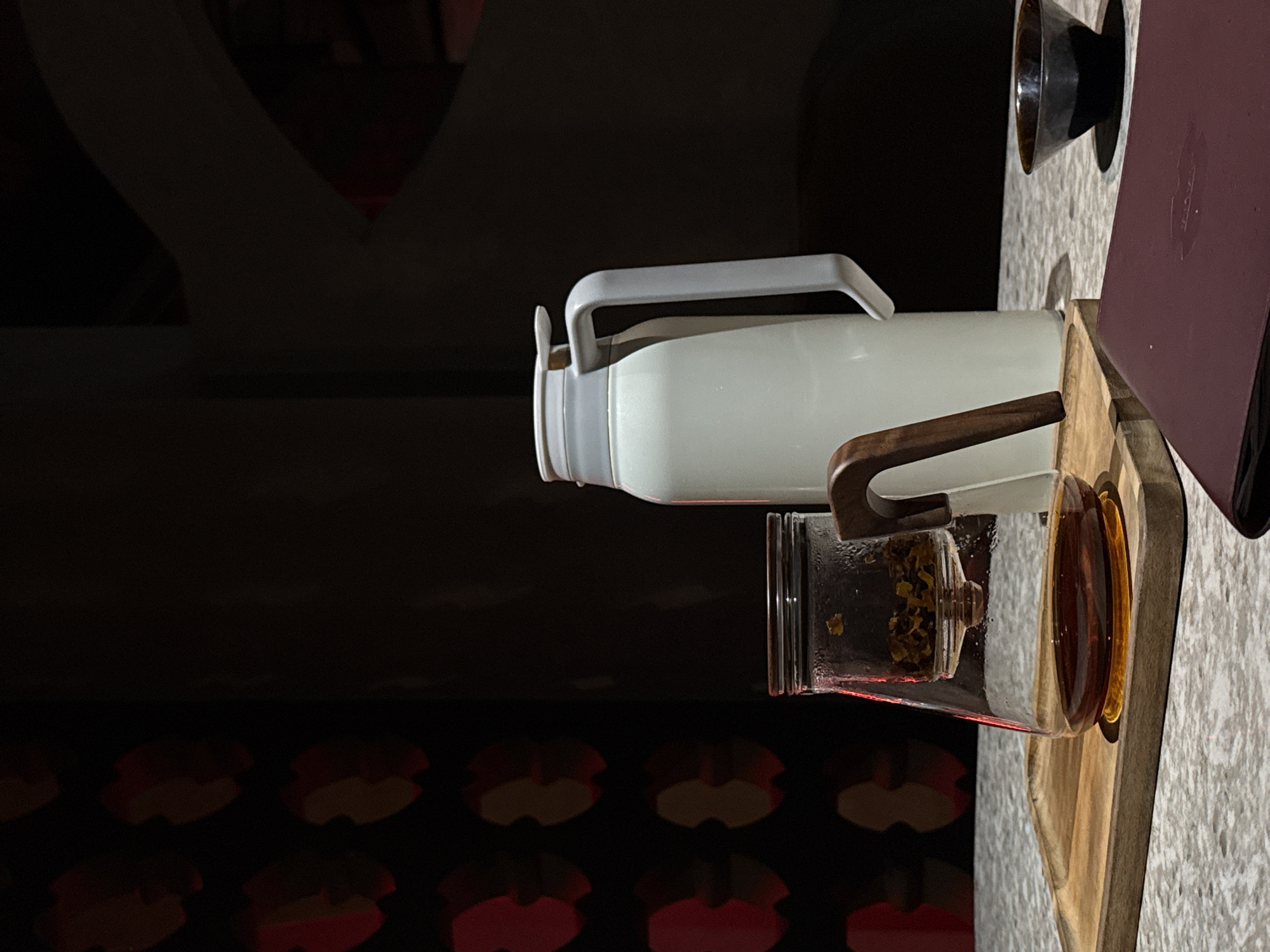 A tea set is arranged on a textured surface, possibly a table in a cafe or restaurant. The setup includes a clear glass teapot with dried tea leaves in the upper infuser and brewed tea in the bottom chamber. Next to it, a tall, white thermos with a grey lid and handle stands prominently. To the right, a small, dark ceramic bowl sits on the surface. The background features a dark wall with a repeating pattern of red and light brown shapes, creating an atmospheric and dimly lit environment. The lighting casts shadows that add depth to the scene. The specific location within the city of Бородино, Russia, is not discernible from the visual information provided. There are no people or animals present in the image, and no actions or interactions are depicted.